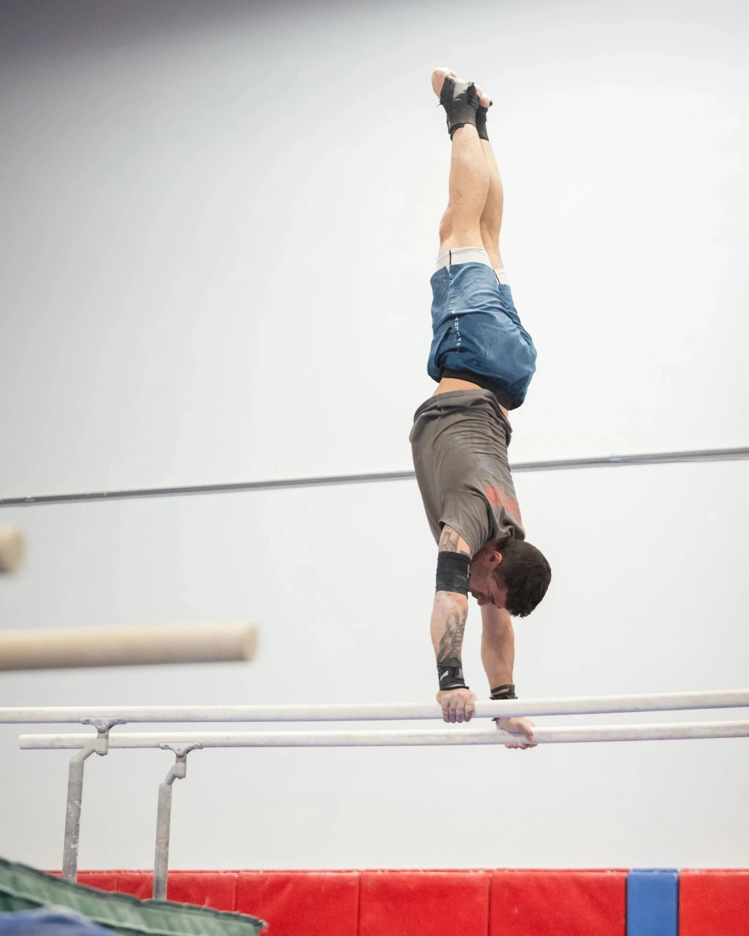 A male gymnast is performing a handstand on parallel bars, wearing a gray T-shirt, shorts, and wrist supports in a gymnasium with a white wall background.