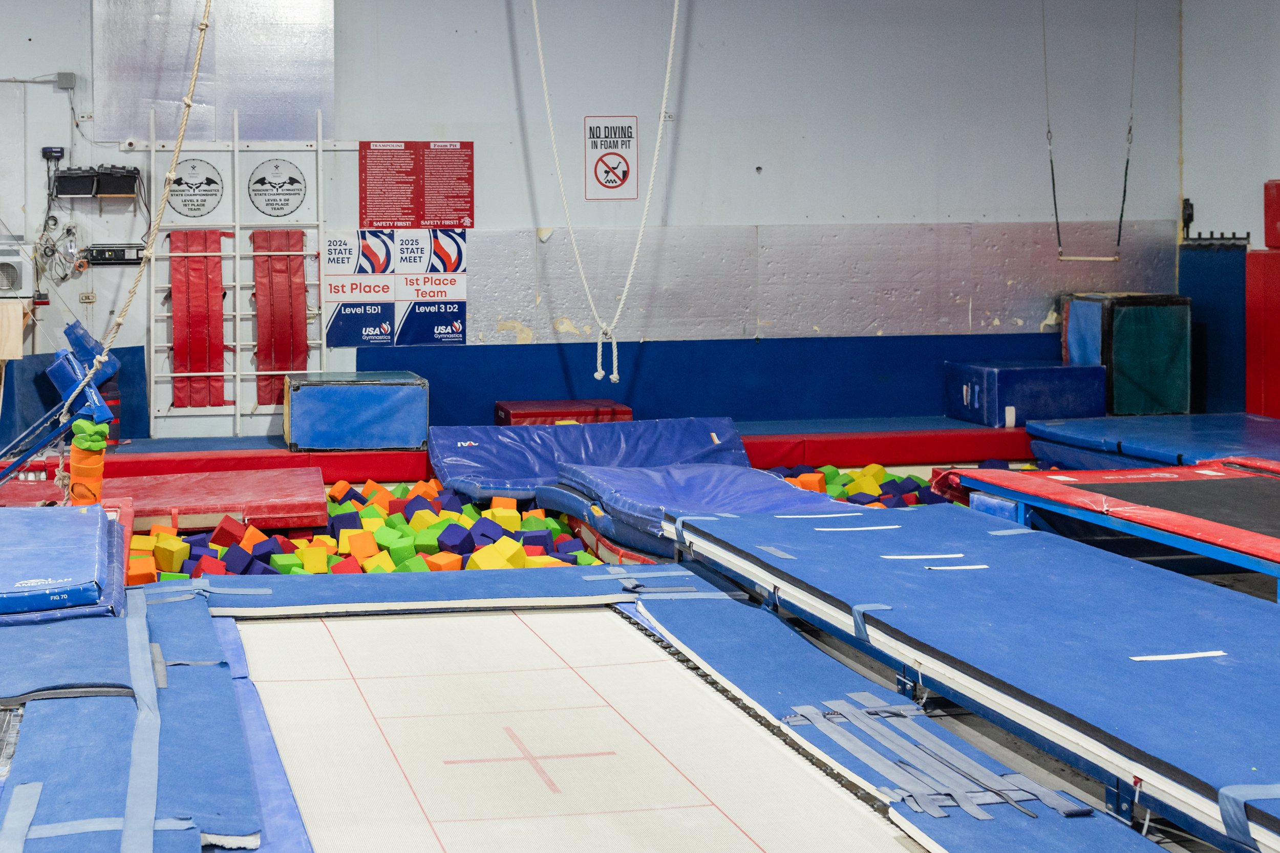 Indoor trampoline gym with foam pit filled with multicolored foam blocks, various trampolines, mats, and safety signs on the wall.