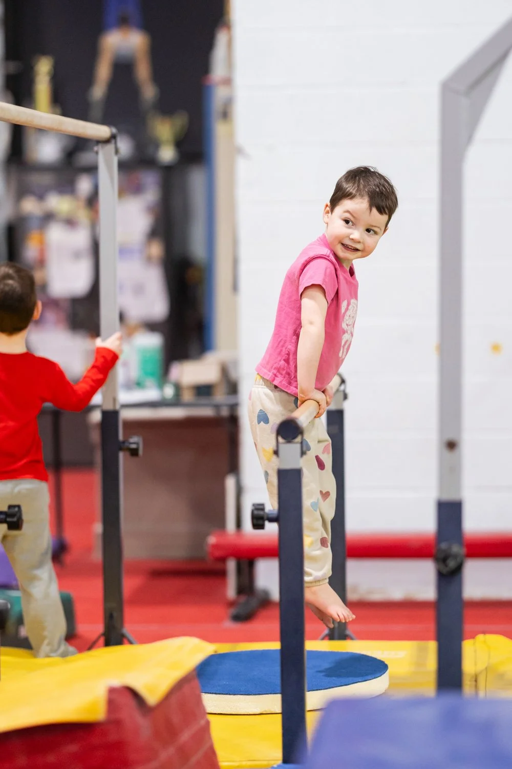 A young  with short dark hair, wearing a pink t-shirt and colorful heart-patterned pants, is balancing on a narrow beam in an indoor gym. She is holding onto the beam with her hands and smiling, with other children and gym equipment visible in the background.