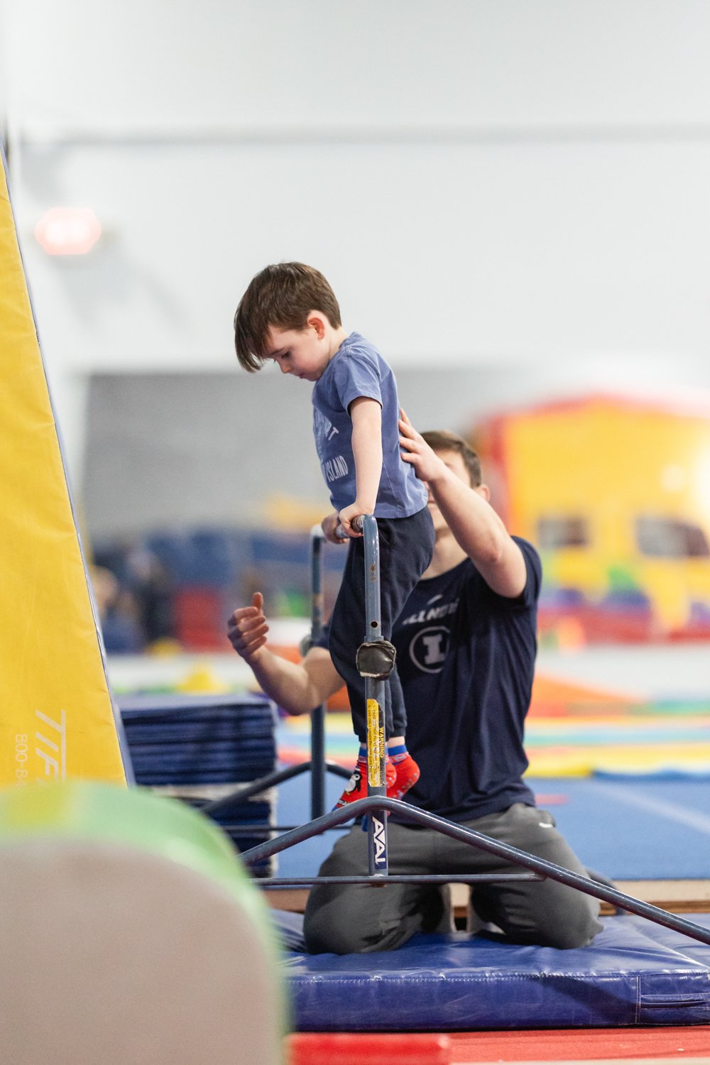 A young boy practicing gymnastics on a pommel horse with assistance from an instructor at an indoor gym.