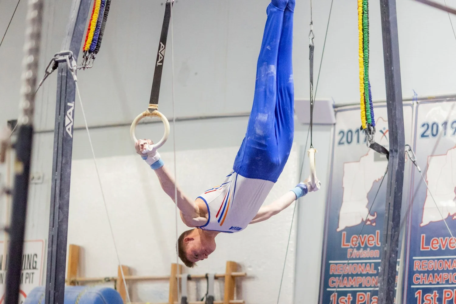 Male gymnast performing on the rings in a gymnastics training facility with banners and equipment in the background.