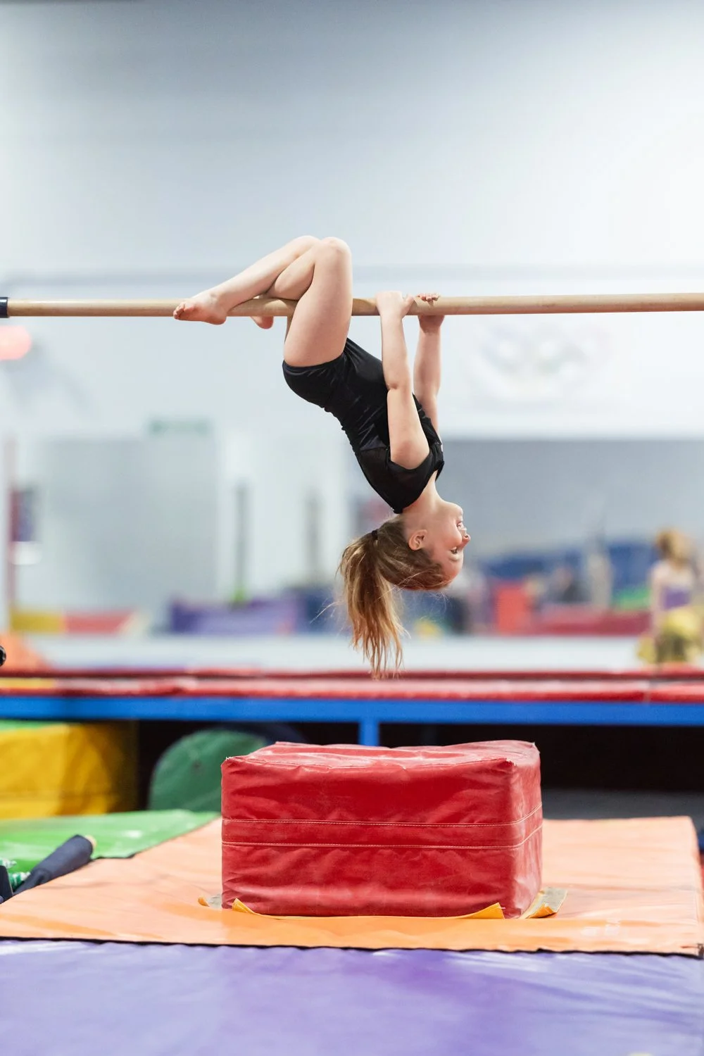 Young girl in black gymnastics outfit hanging upside down on a horizontal bar in a gymnasium, with colorful mats and equipment in the background.