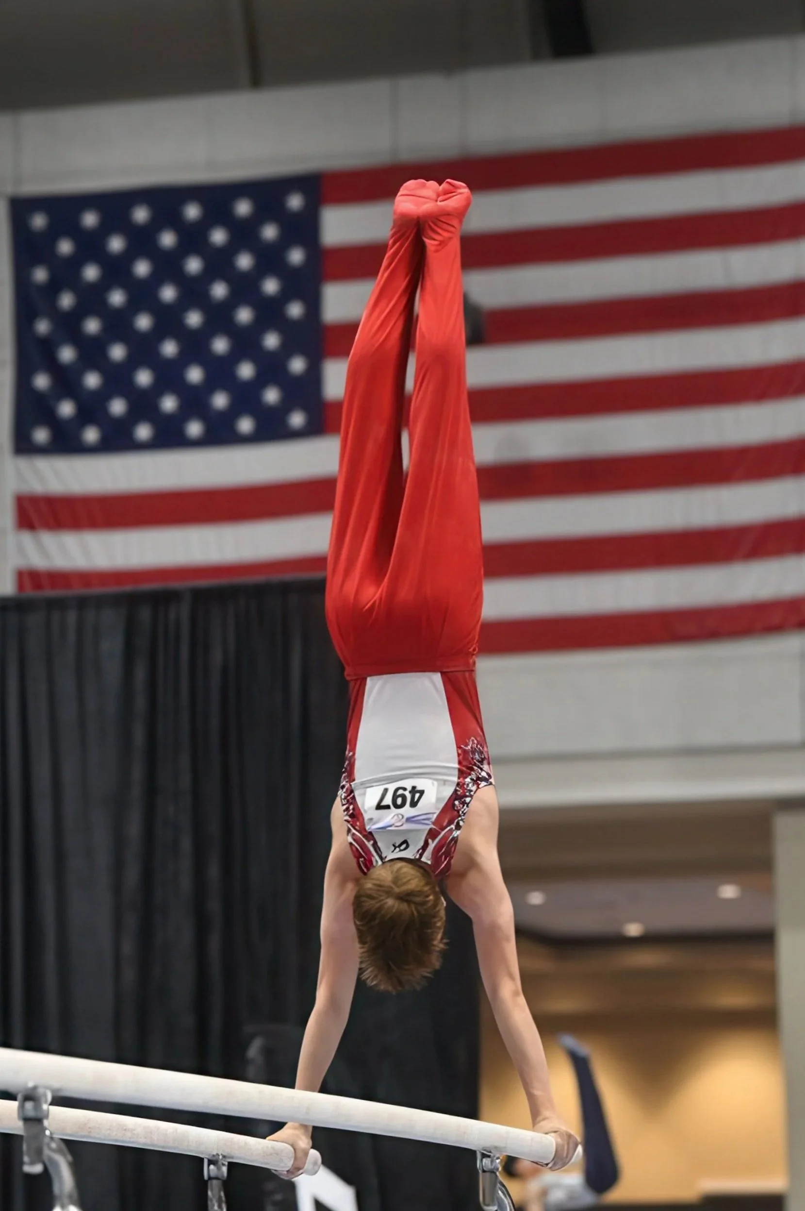 A gymnast performing a handstand on the balance beam at a competition, with an American flag hanging in the background.