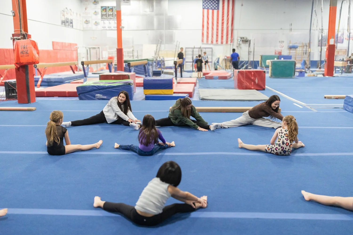 Children and instructors stretching on a blue gymnastics mat in a gymnastics training facility.