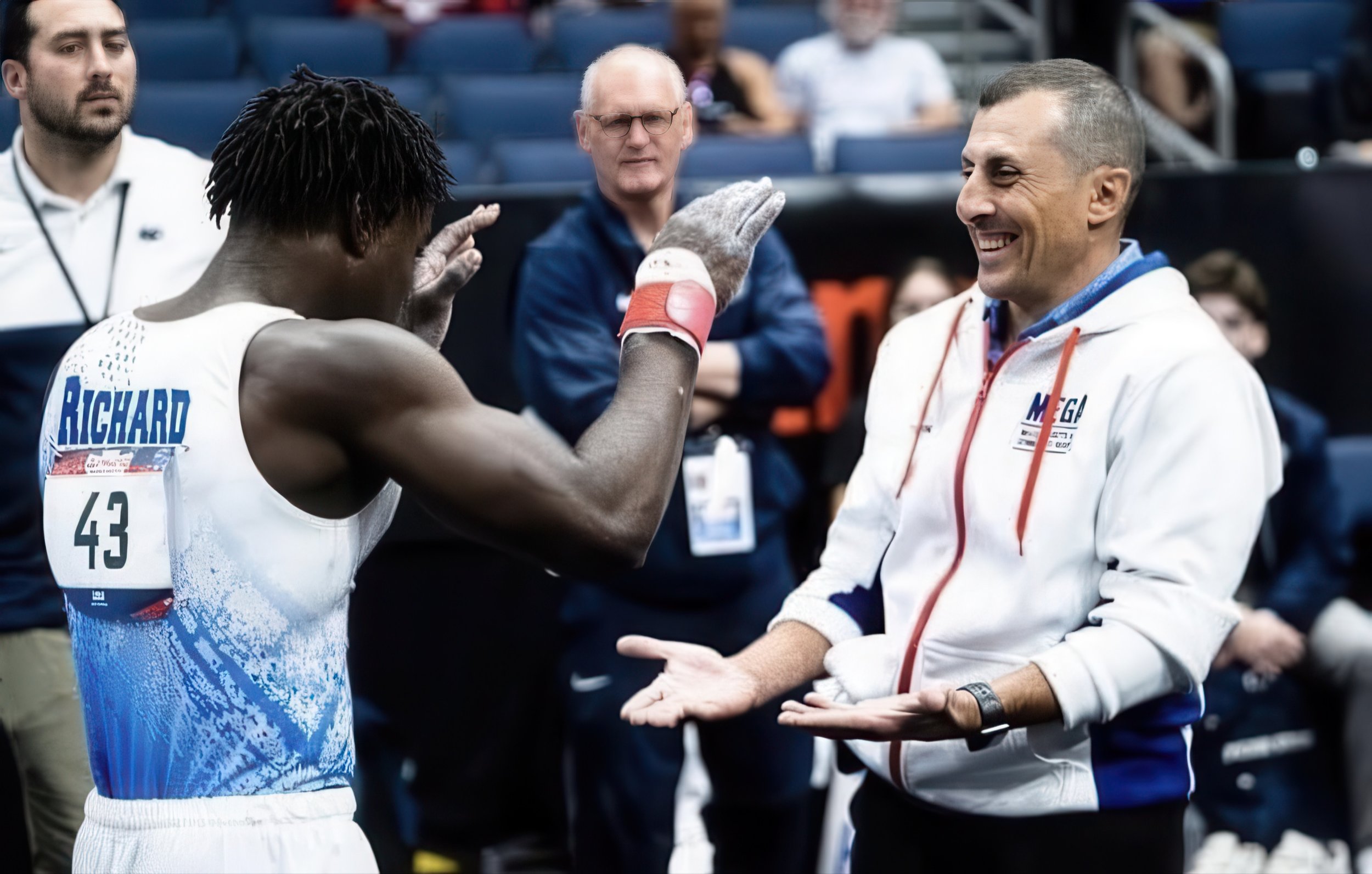 A male athlete with the name Richard on his jersey is having a conversation with a man wearing a white jacket with the MEGA logo at a sports event. The athlete is gesturing with his hands, and the other man has his palms up in a questioning manner. There are other people in the background watching.