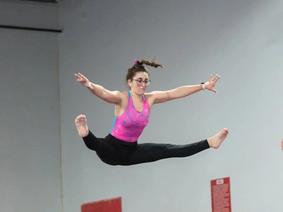 Young woman in athletic clothing performing a split leap in a gym.