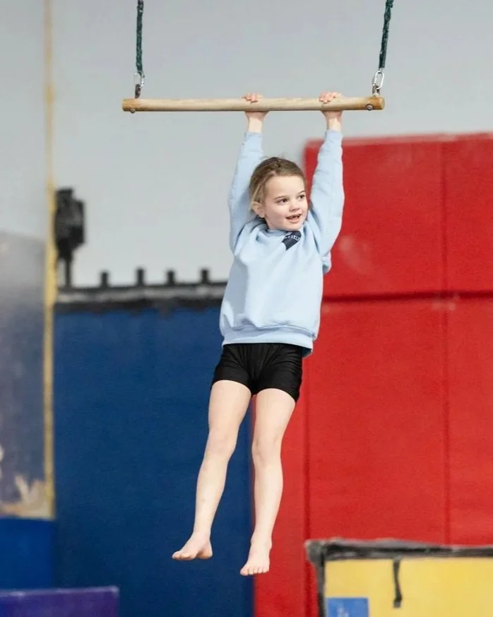 A young girl in a light blue hoodie and black shorts hanging from a gymnastics bar in a gymnasium.