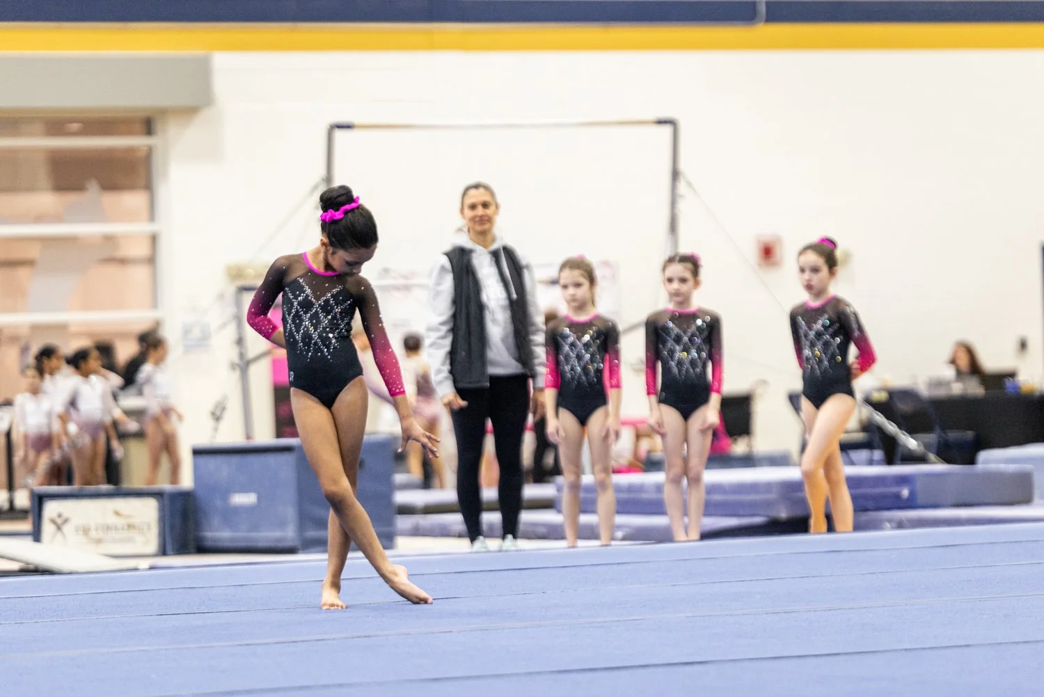 Young female gymnast in a black and pink leotard practicing a routine on blue gymnastics mat, with her coach and other gymnasts observing in the background at an indoor gym.