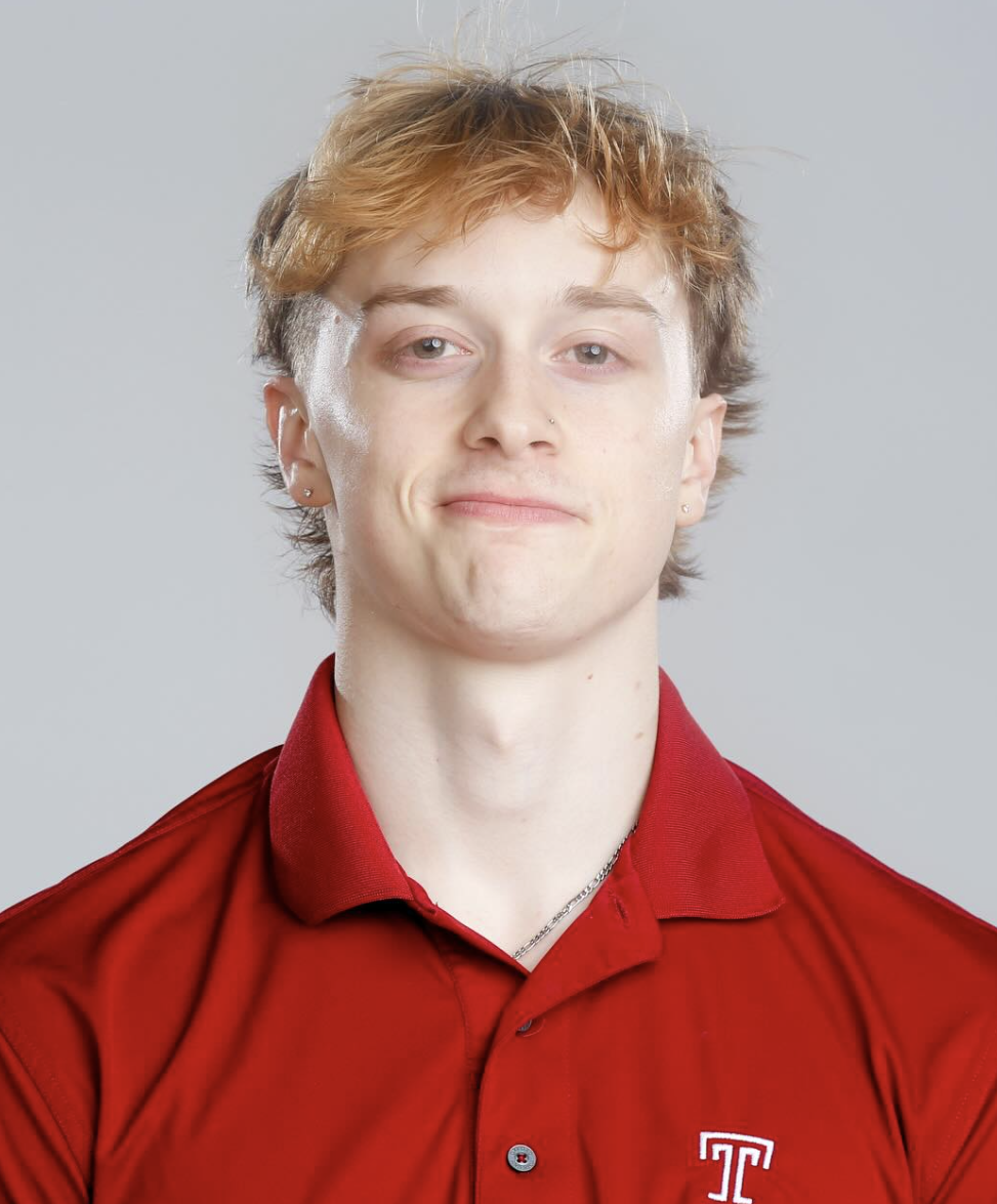 A young person with short, wavy, blond hair, fair skin, wearing a red collared shirt with a white and red logo on the chest, and small earrings, posing against a plain light gray background.