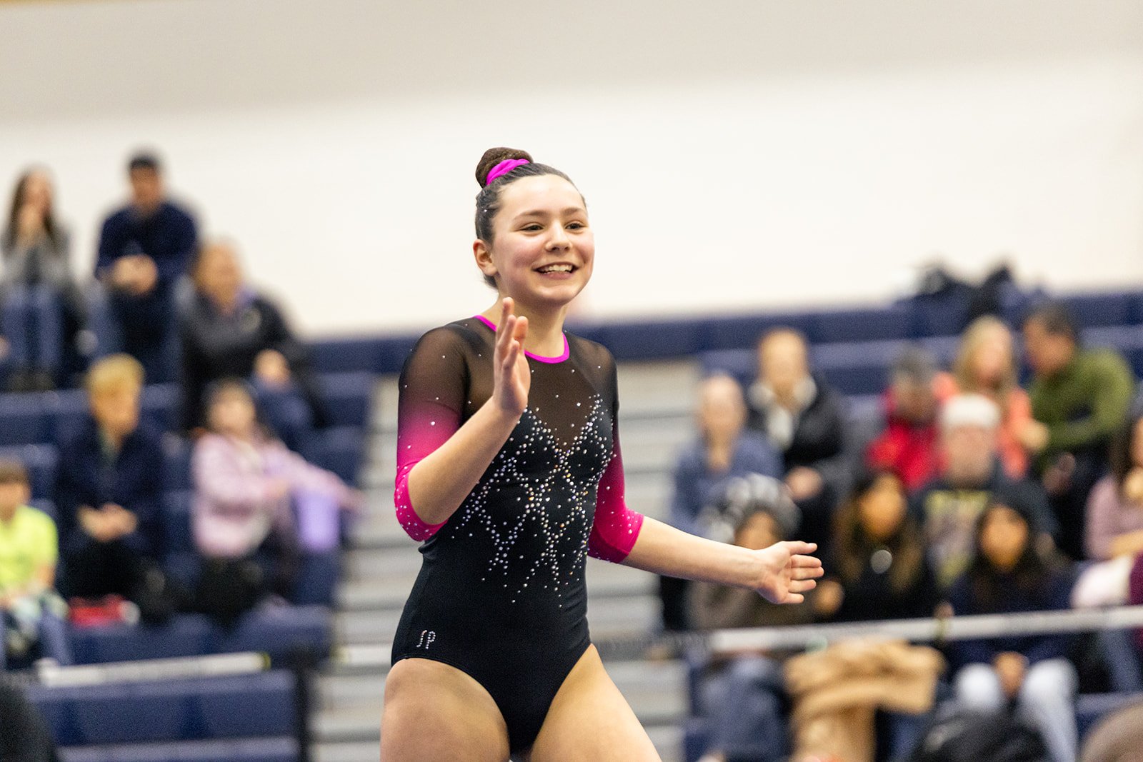 A young female gymnast smiling and waving in a black and pink leotard during a gymnastics event, with spectators in the background.