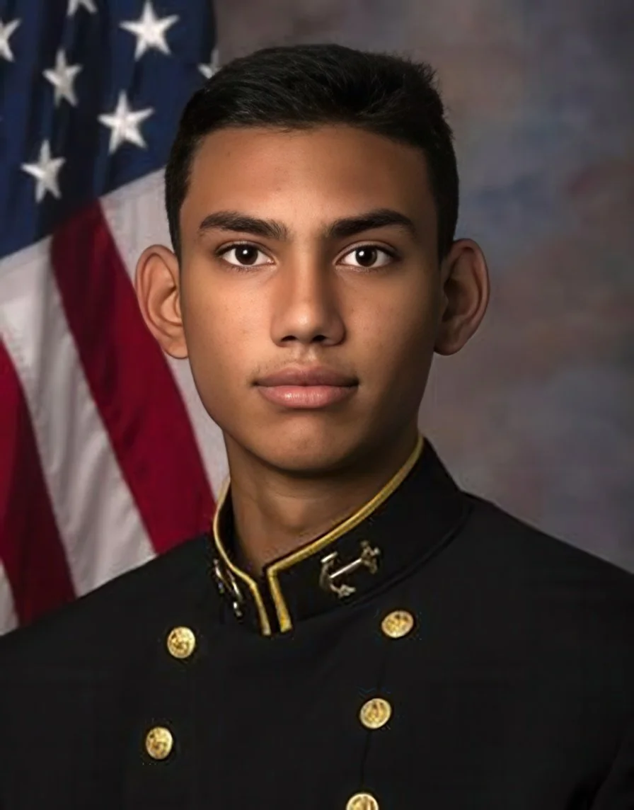 A young man in a black military uniform with gold accents, standing in front of an American flag.