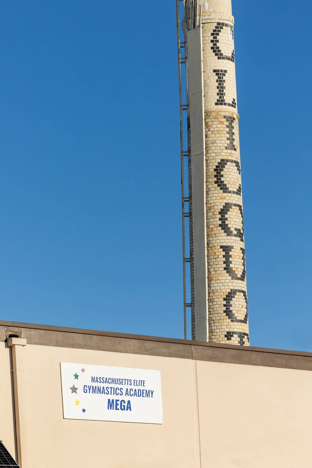 A tall brick chimney with the words 'GRO' spelled vertically in black mosaic tiles, and part of a sign reading 'Massachusetts Elite Gymnastics Academy Mega' on a beige wall, under a clear blue sky.