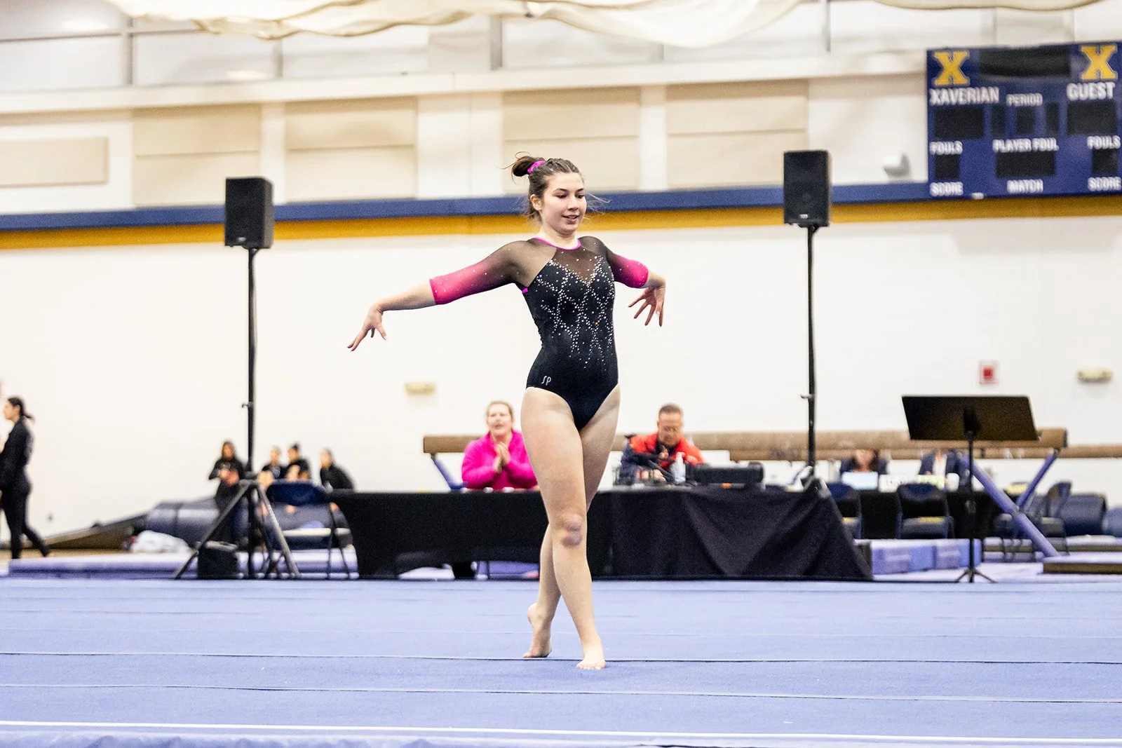 A young female gymnast performing a routine on a blue gymnastics floor, wearing a black and pink leotard, with judges and spectators in the background.