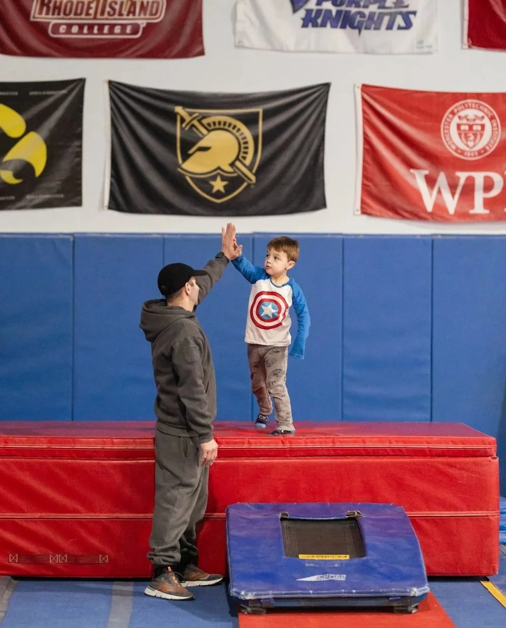A young boy dressed in a Captain America shirt high-fives a man at a gymnastics facility. The boy stands on a red padded platform with blue wall padding behind him, and flags hang on the wall above.