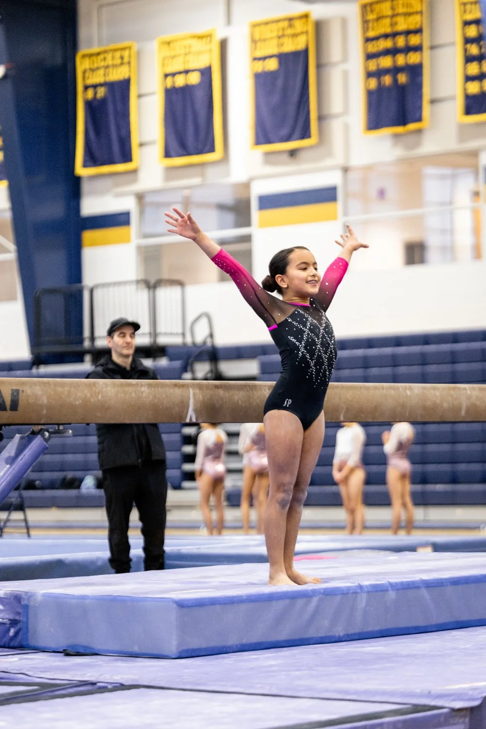 Young gymnast in a black and pink leotard celebrating after performing on the balance beam in an indoor gymnastics facility, with other gymnasts and a judge or coach in the background.