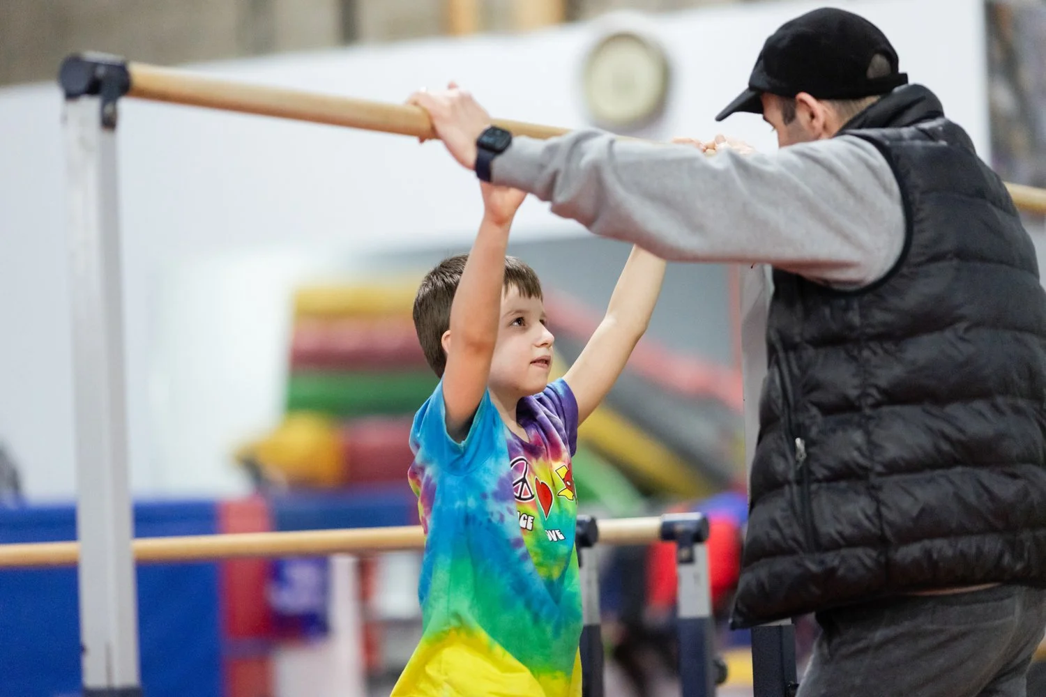 A young boy in a colorful tie-dye shirt practicing gymnastics or dance with the assistance of an instructor in a gym.