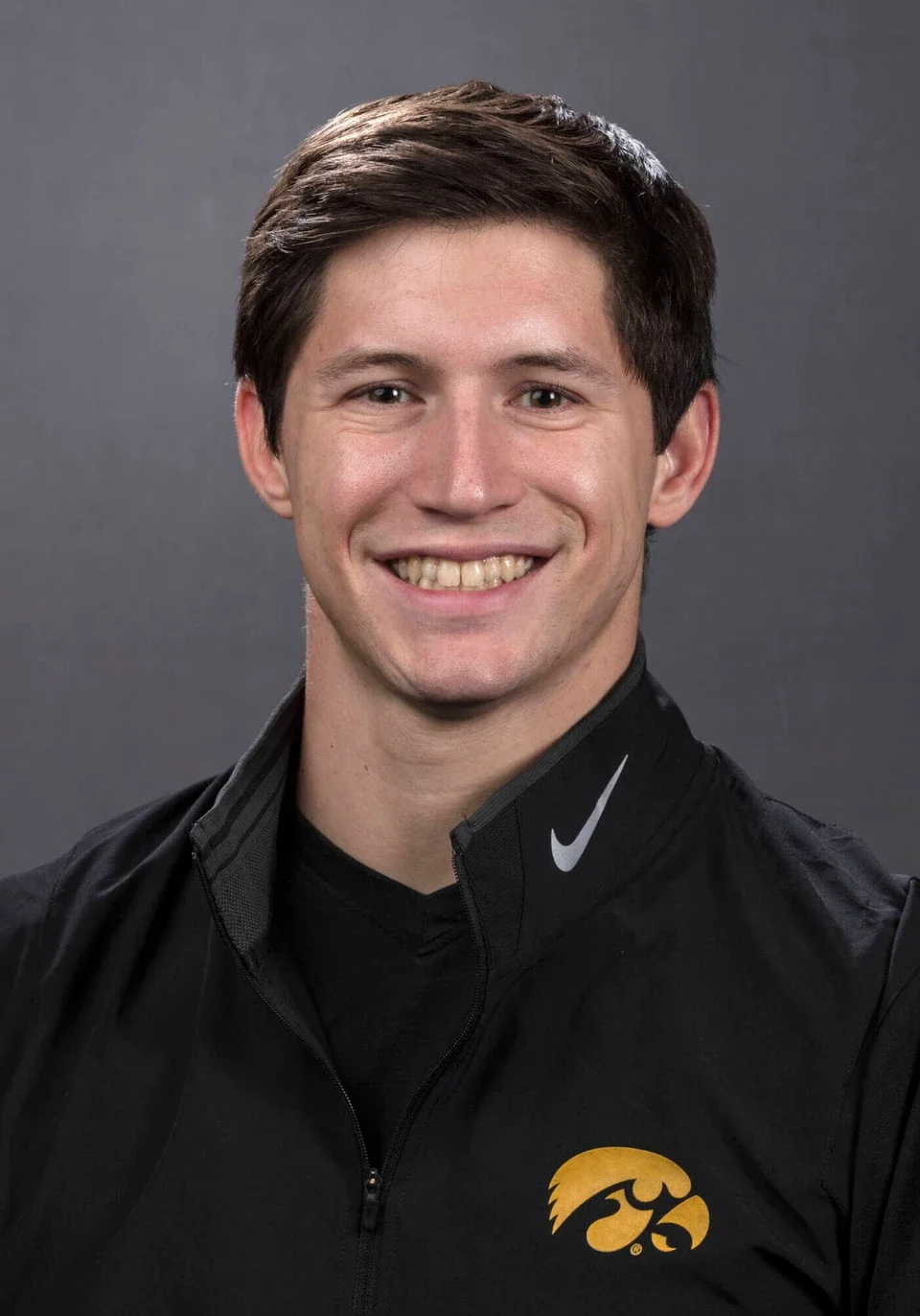 Young man smiling, wearing black athletic jacket with Nike logo and Iowa Hawkeyes logo, against a gray background.
