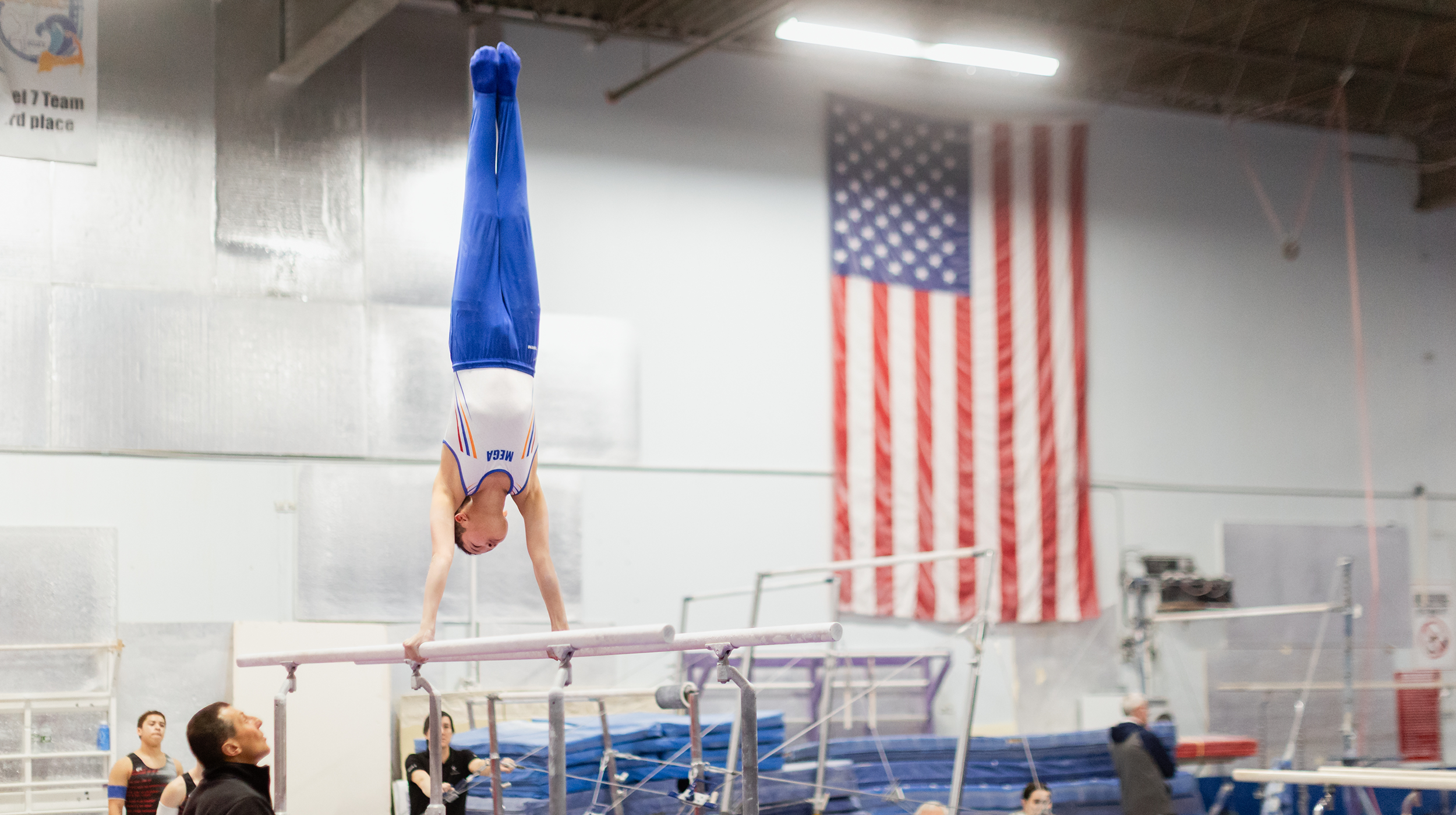 A gymnast performing a handstand on the balance beam in a gymnasium with an American flag in the background.