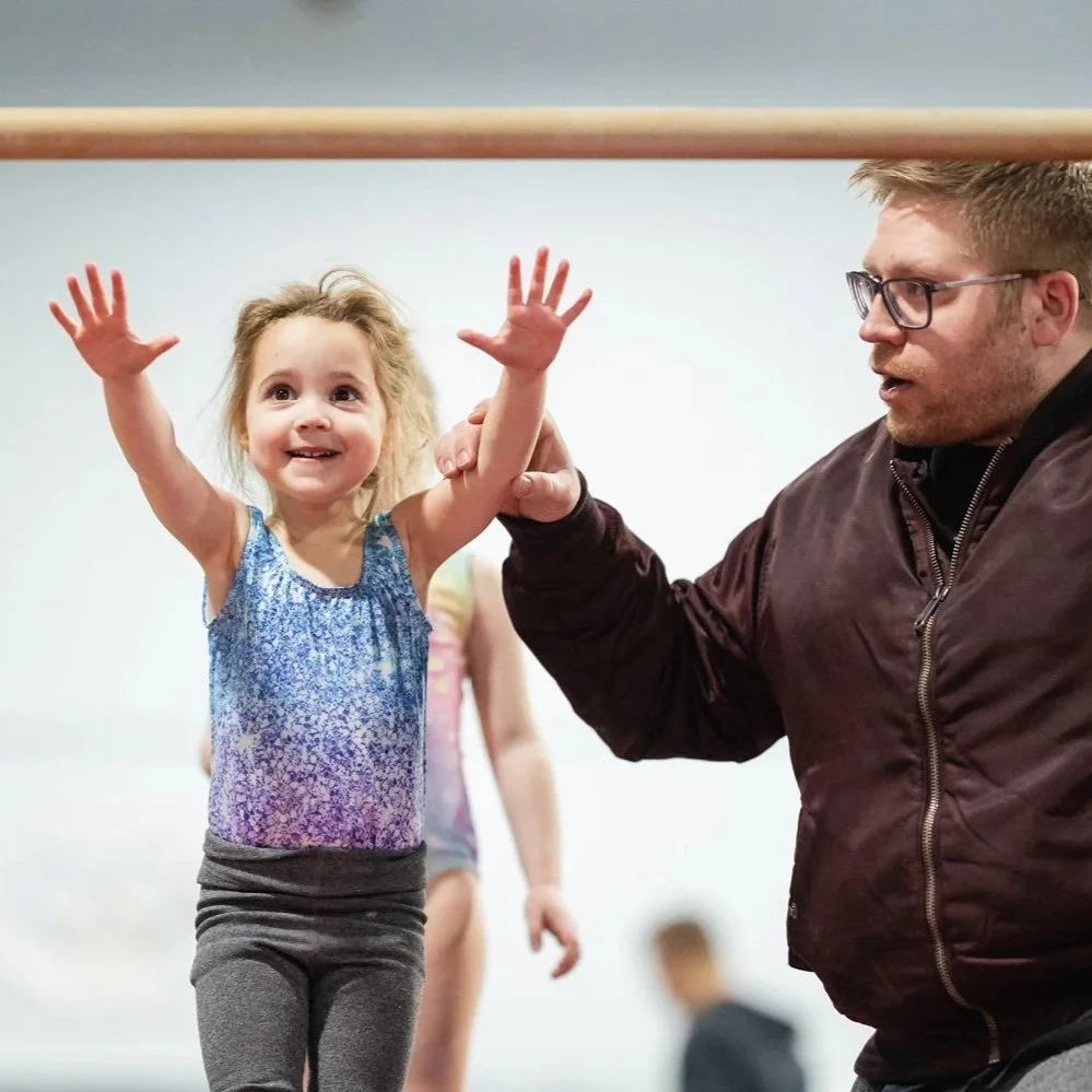 A young girl with curly blonde hair and a blue patterned tank top smiling and raising her arms while an coach supports her.