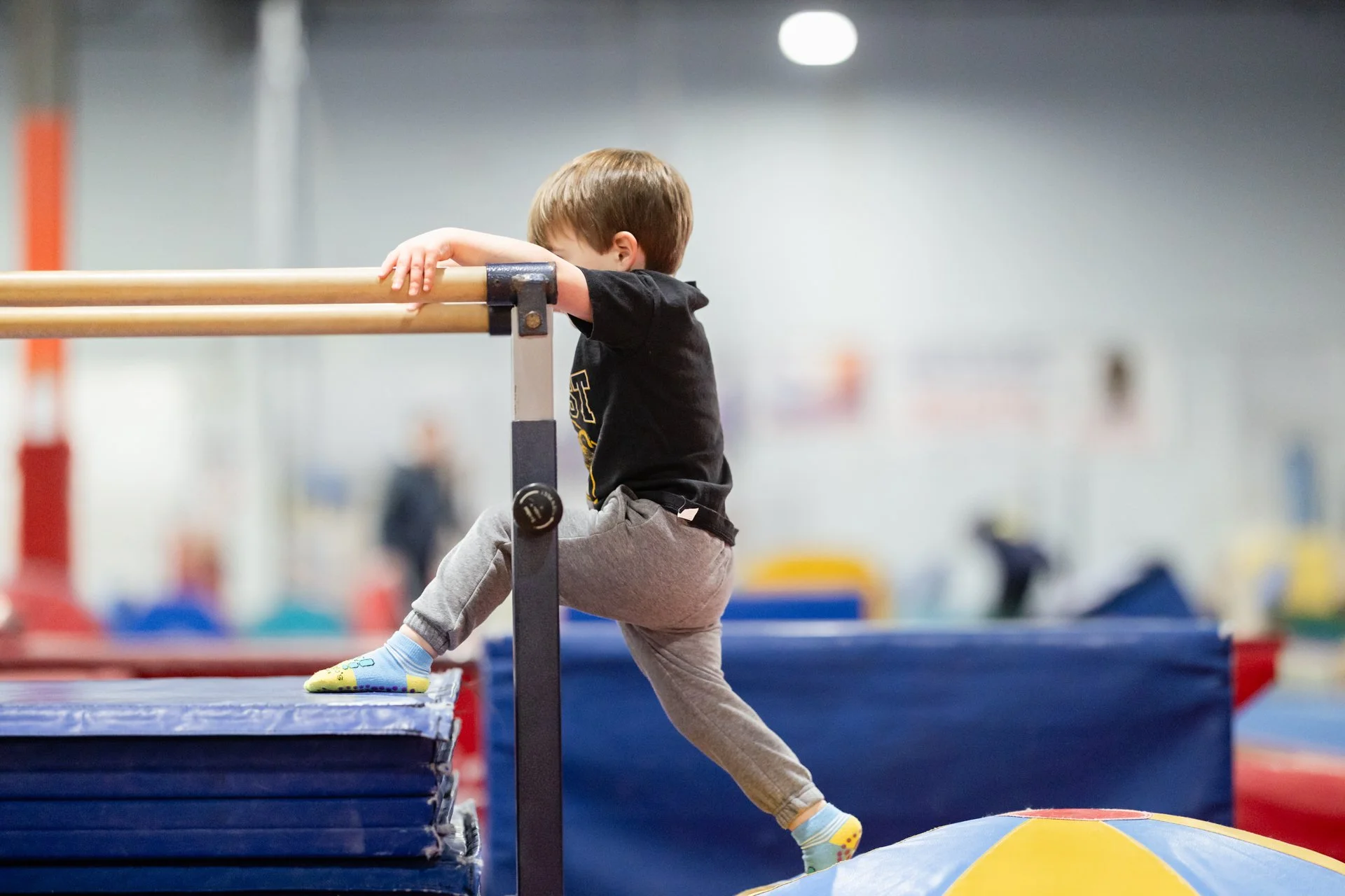 A young boy wearing a black shirt and gray pants is hanging from a ballet barre in an indoor gym. He is gripping the bar with both hands and lifting his legs, with his face turned down and to the side. The background is blurry with gym equipment and colorful mats.