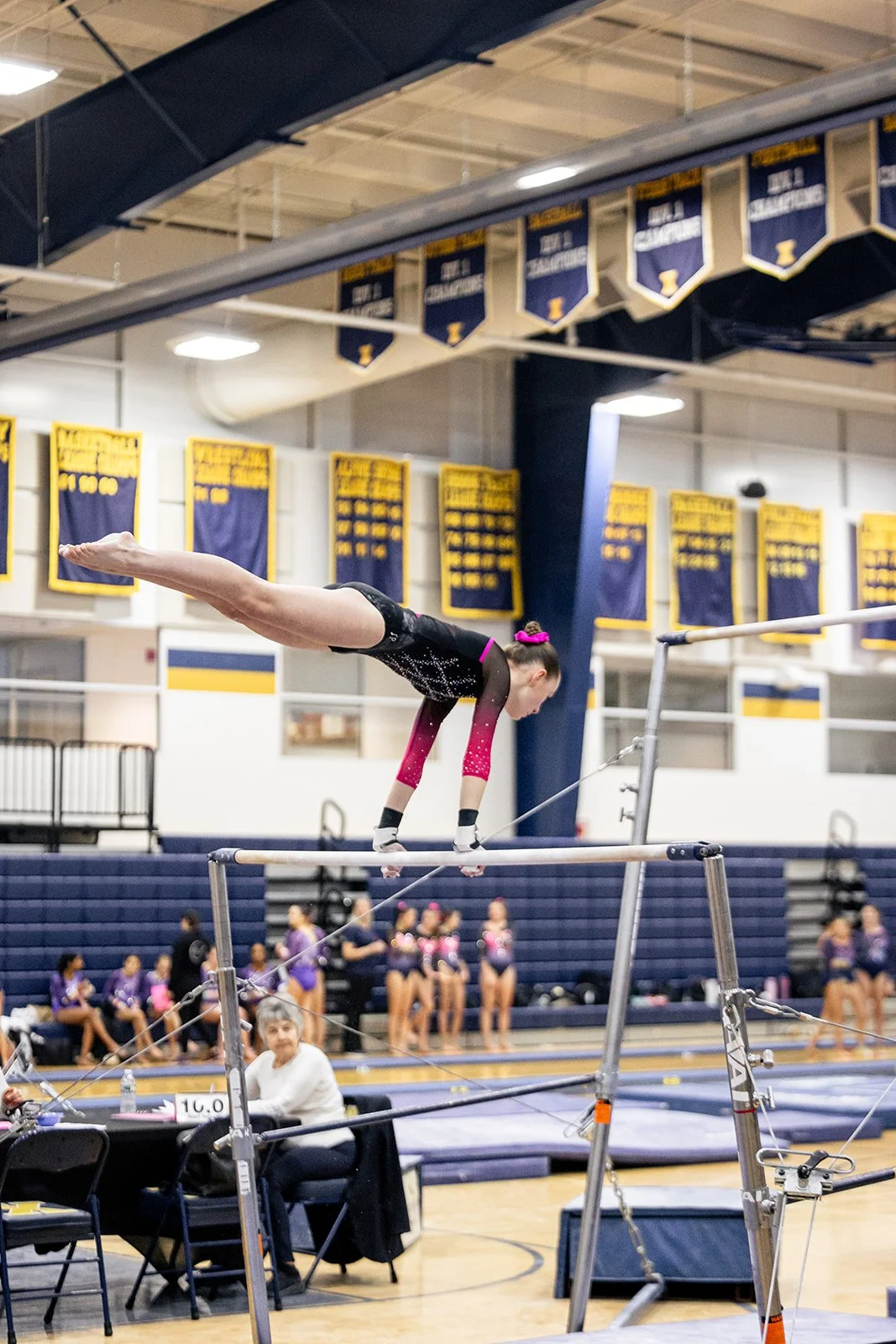 Gymnast performing on the balance beam during a gymnastics meet with banners hanging in the background.