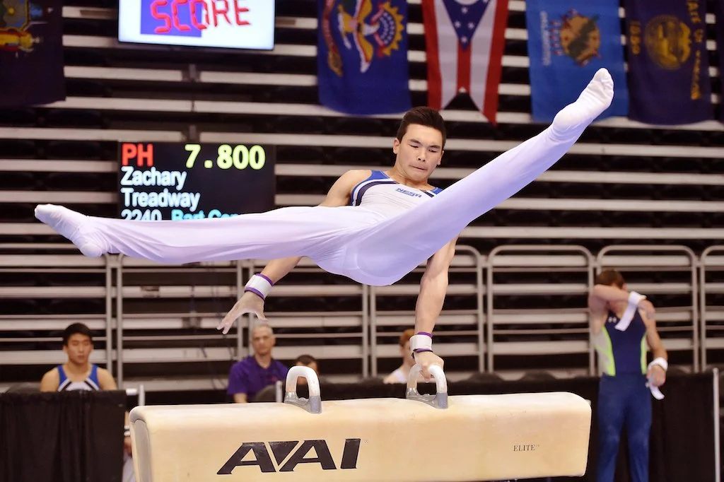 Male gymnast in white attire performing a routine on the pommel horse during a gymnastics competition.
