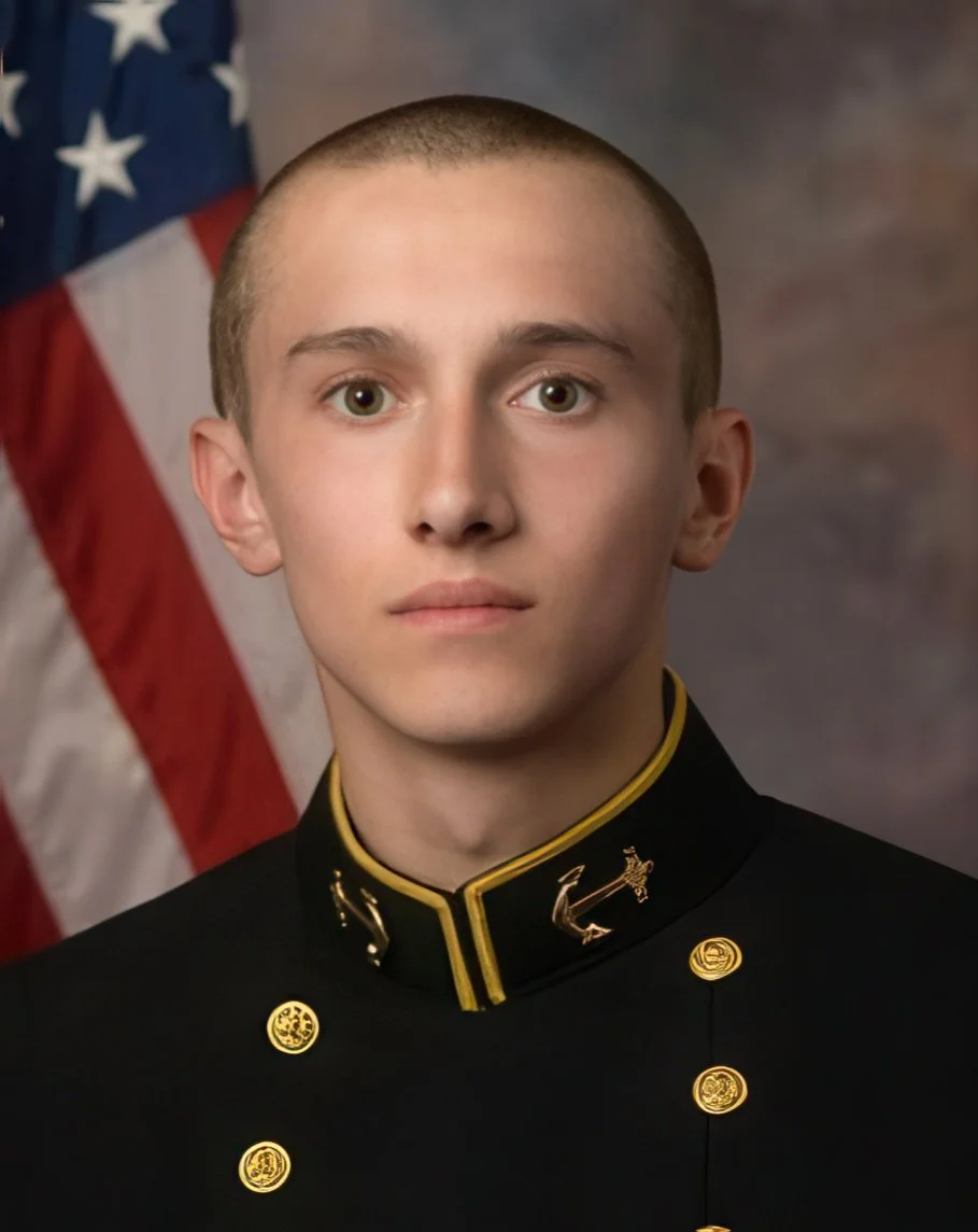 Young man in a military dress uniform with an American flag in the background, looking directly at the camera.