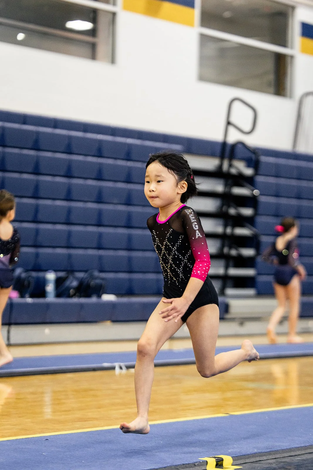 Young girl in black and pink gymnastics leotard doing a running warm-up on a gym mat inside a gymnasium, with other gymnasts in the background.