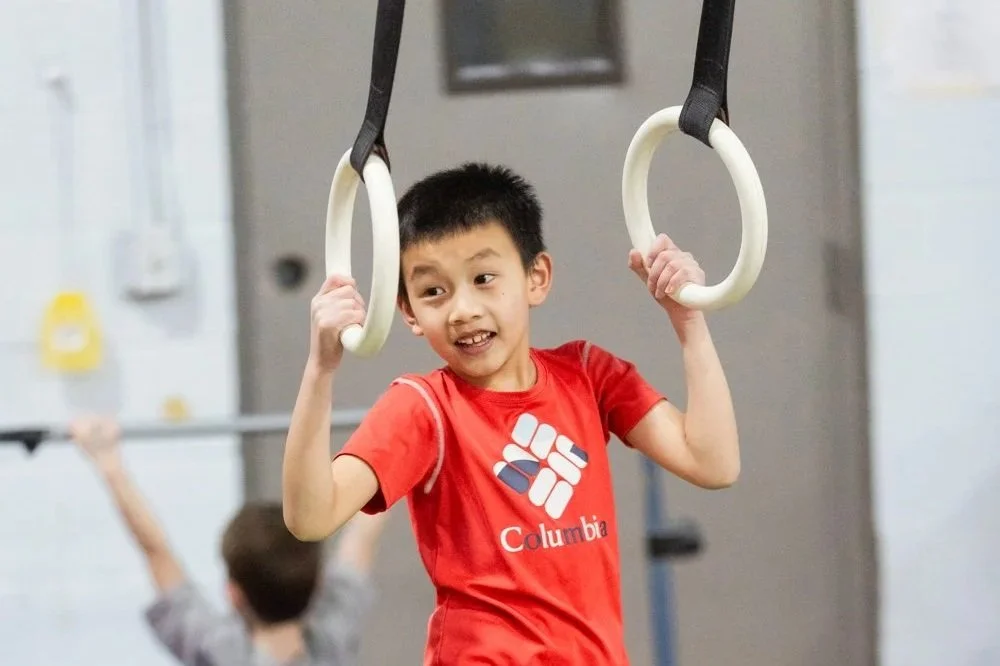 A young boy in a red Columbia t-shirt hanging from gymnastic rings in a gym or fitness center.