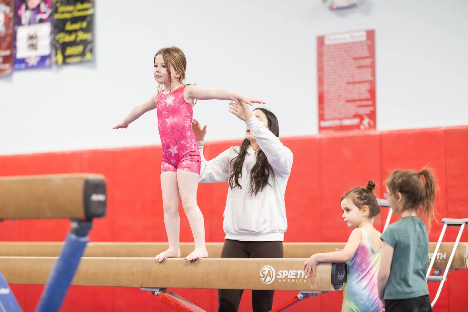 A young girl in a pink leotard performs a balance on a gymnastics beam, with supervision from an adult coach and two other children watching nearby in a gymnastics gym.