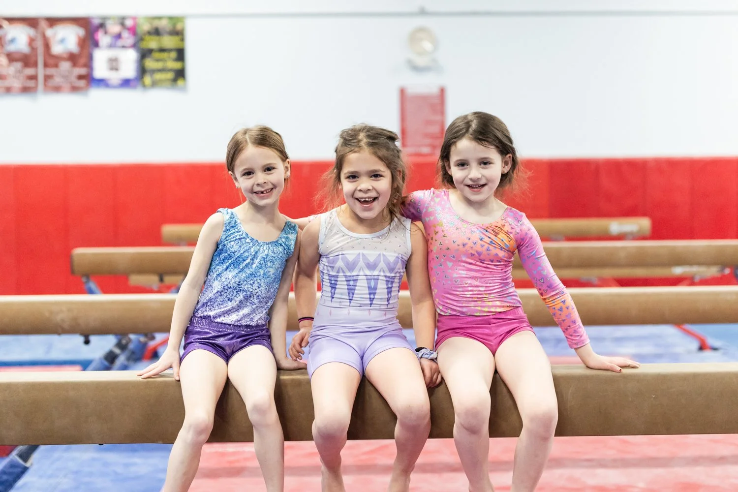 Three young girls in colorful gymnastics outfits sitting on a balance beam in a gymnasium, smiling at the camera.