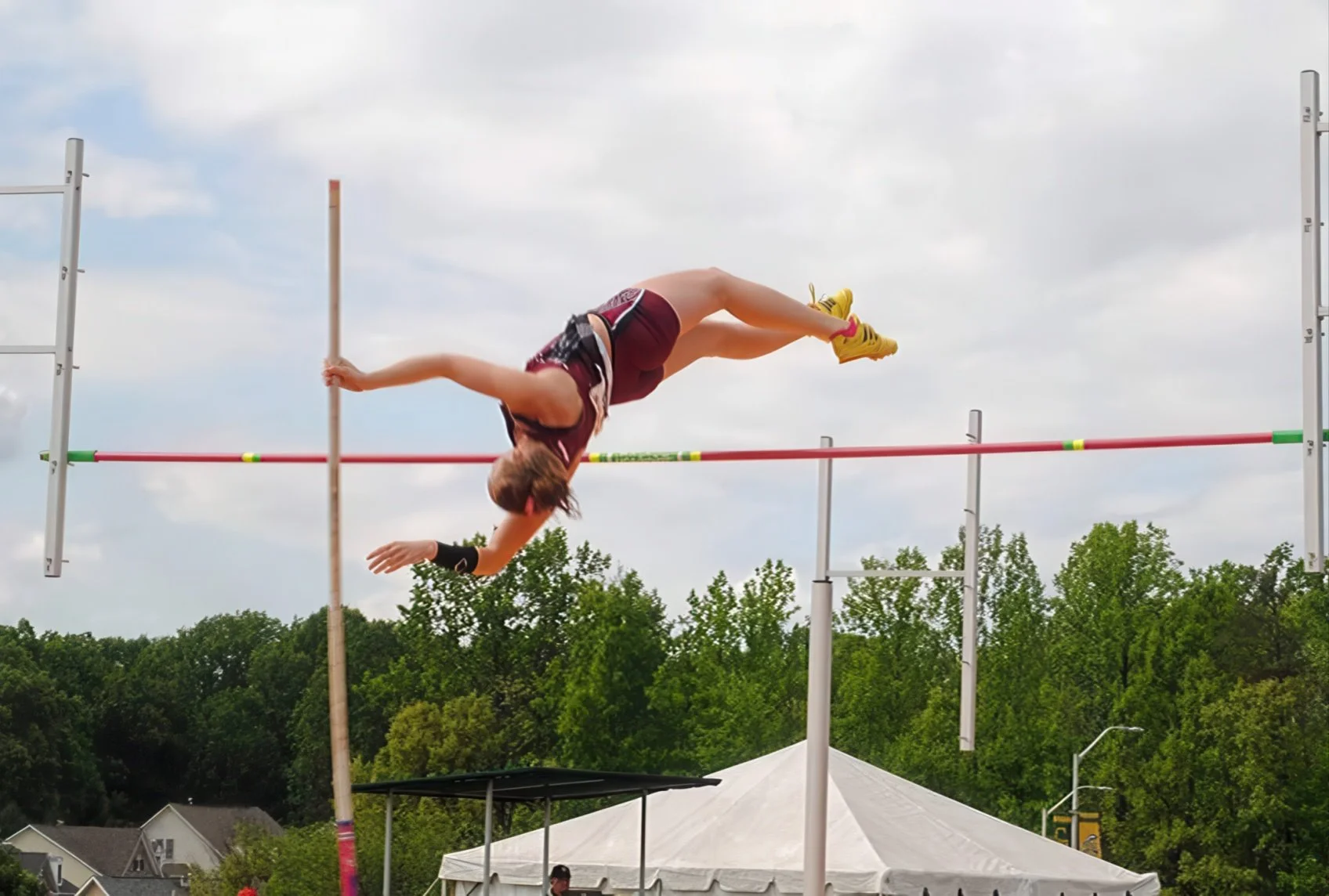 A female athlete in a maroon sports outfit is performing a pole vault over a horizontal bar outdoors with green trees, houses, and a tent in the background.