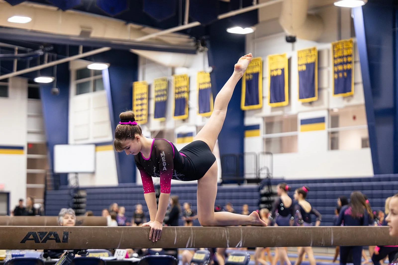 Young female gymnast in black and pink leotard performing a balance move on the balance beam in a gymnasium with banners hanging on the wall and spectators in the background.