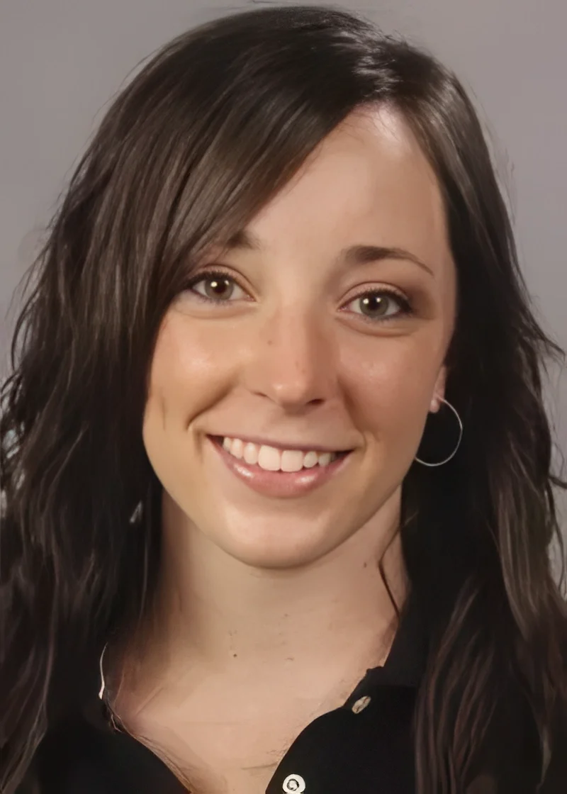 A young woman with long, wavy dark brown hair, light green eyes, and earrings, smiling at the camera against a neutral background.