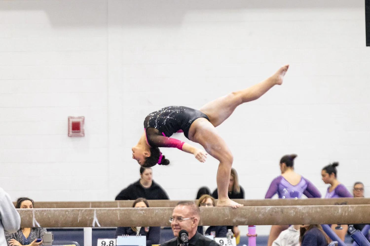 A female gymnast is performing a handstand on a balancing beam during a gymnastics competition, with judges and spectators watching.