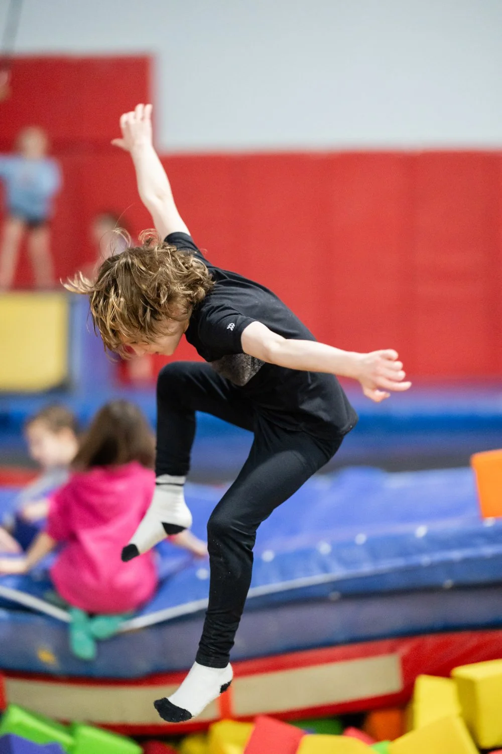A young boy with curly hair is mid-air, falling or jumping at an indoor trampoline park with foam blocks in the foreground and other children in the background.