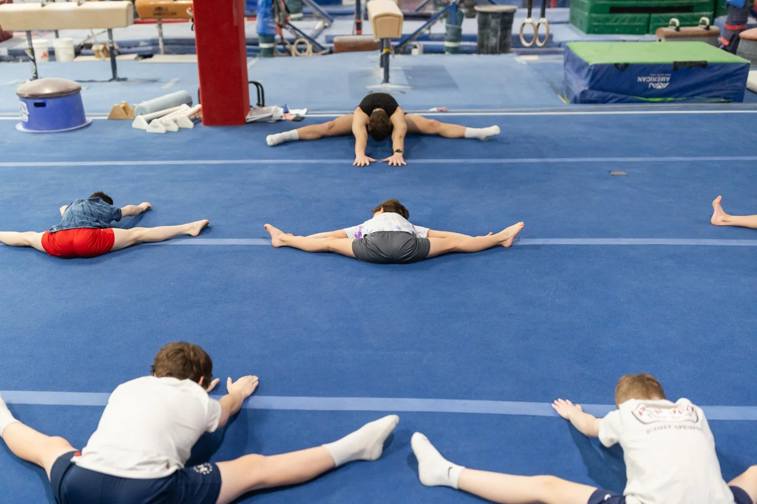 Children and an adult practicing split stretches on a blue gymnastics mat during a gymnastics class or training session.