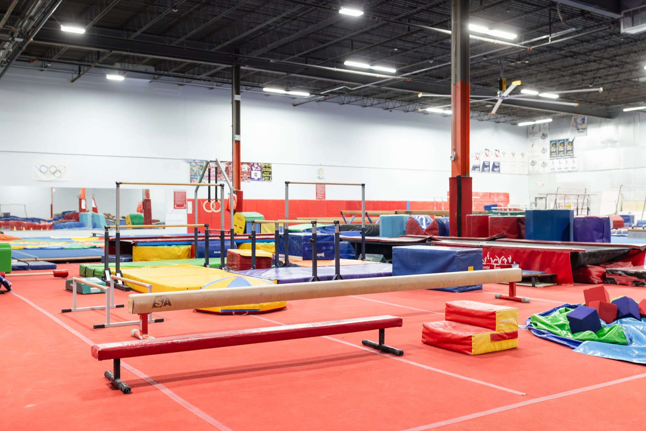 An indoor gymnastics training facility with various gymnastics equipment, mats, and padding on a red floor, white walls, and a high ceiling with industrial lighting and ceiling fans.
