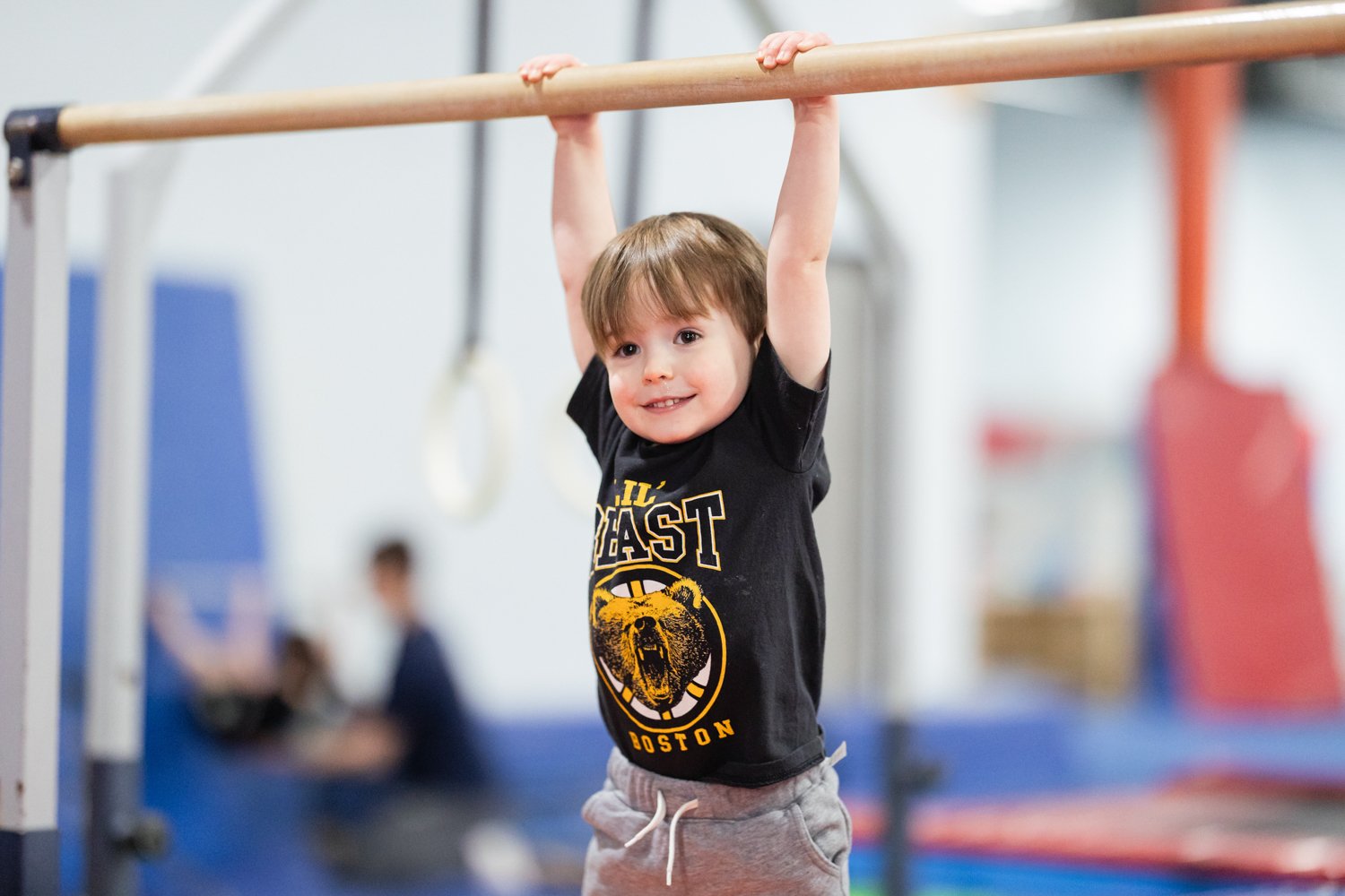 A young boy hanging from a gymnastics bar in a gym. He is smiling and wearing a black shirt with a bear and the words 'ALL BEAST BOSTON.' In the background, another person is sitting on a blue padded mat with blurred gym equipment.