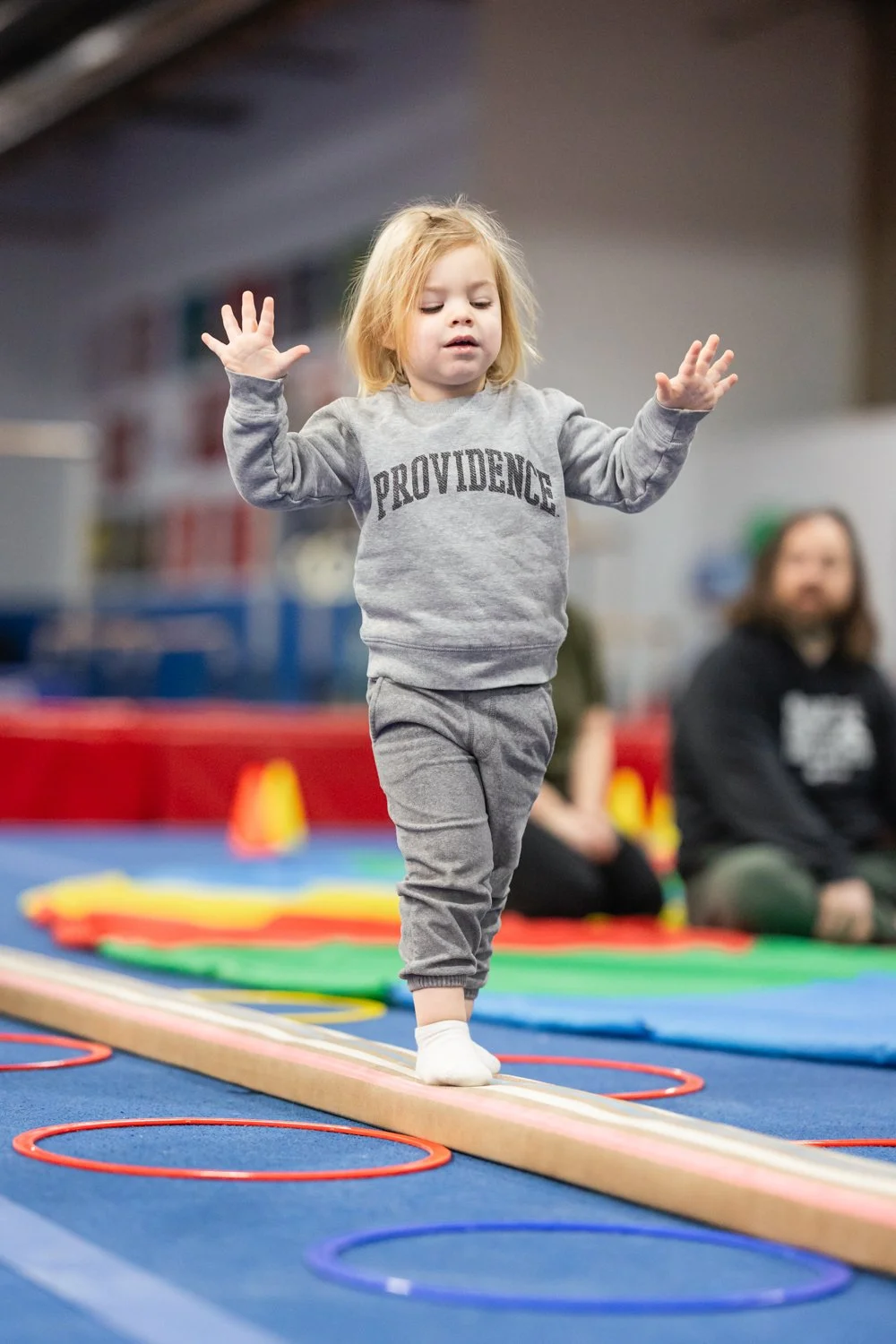 A young girl balancing on a wooden beam with her arms raised during an indoor gym activity, with colorful hoops on the floor and other children and adults in the background.