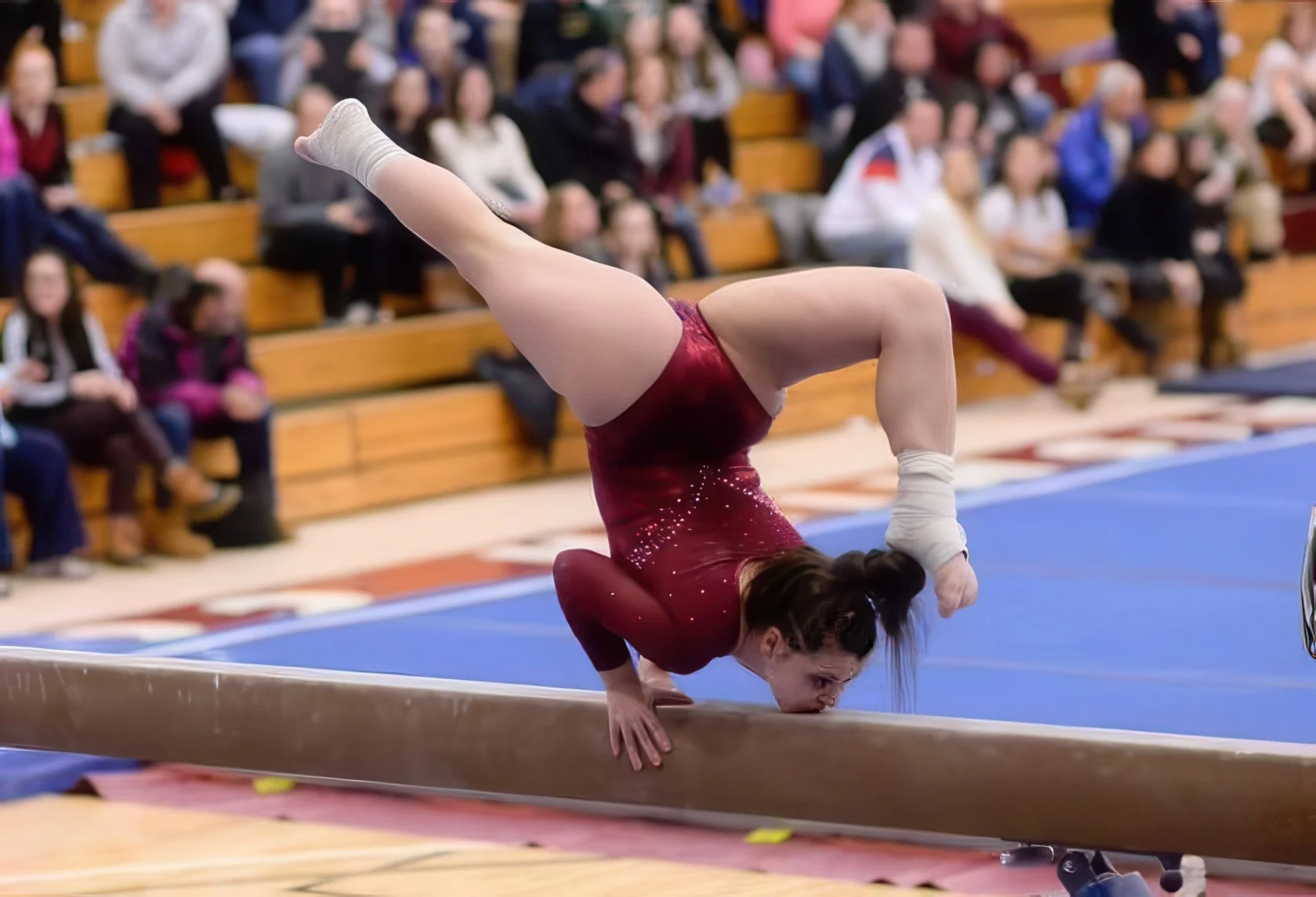 A female gymnast in a red leotard performs a move on the balance beam during a gymnastics competition, with an audience seated in bleachers watching.