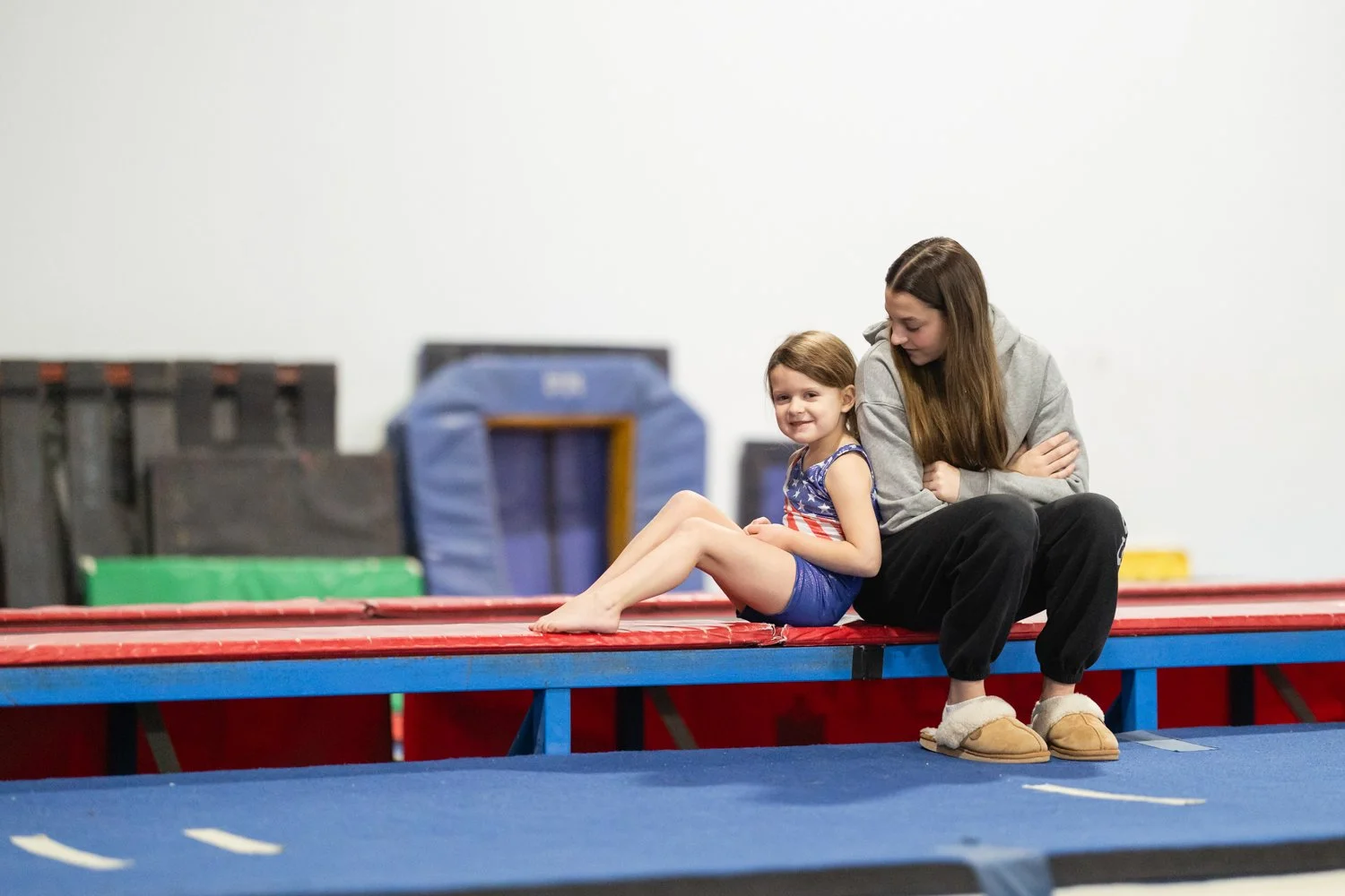 A young girl and her coach sit together on a gymnastics trampoline mat in a gym, smiling and looking at the camera.