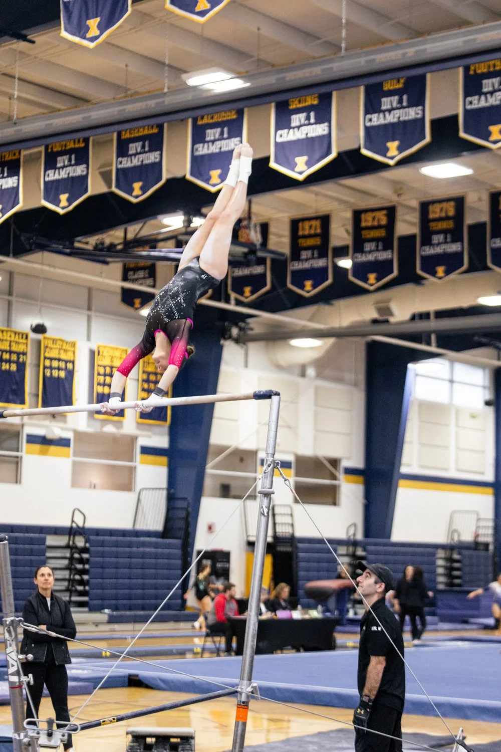 A gymnast performing a handstand on the uneven bars during a gymnastics meet inside a gymnasium, with coaches and judges watching.