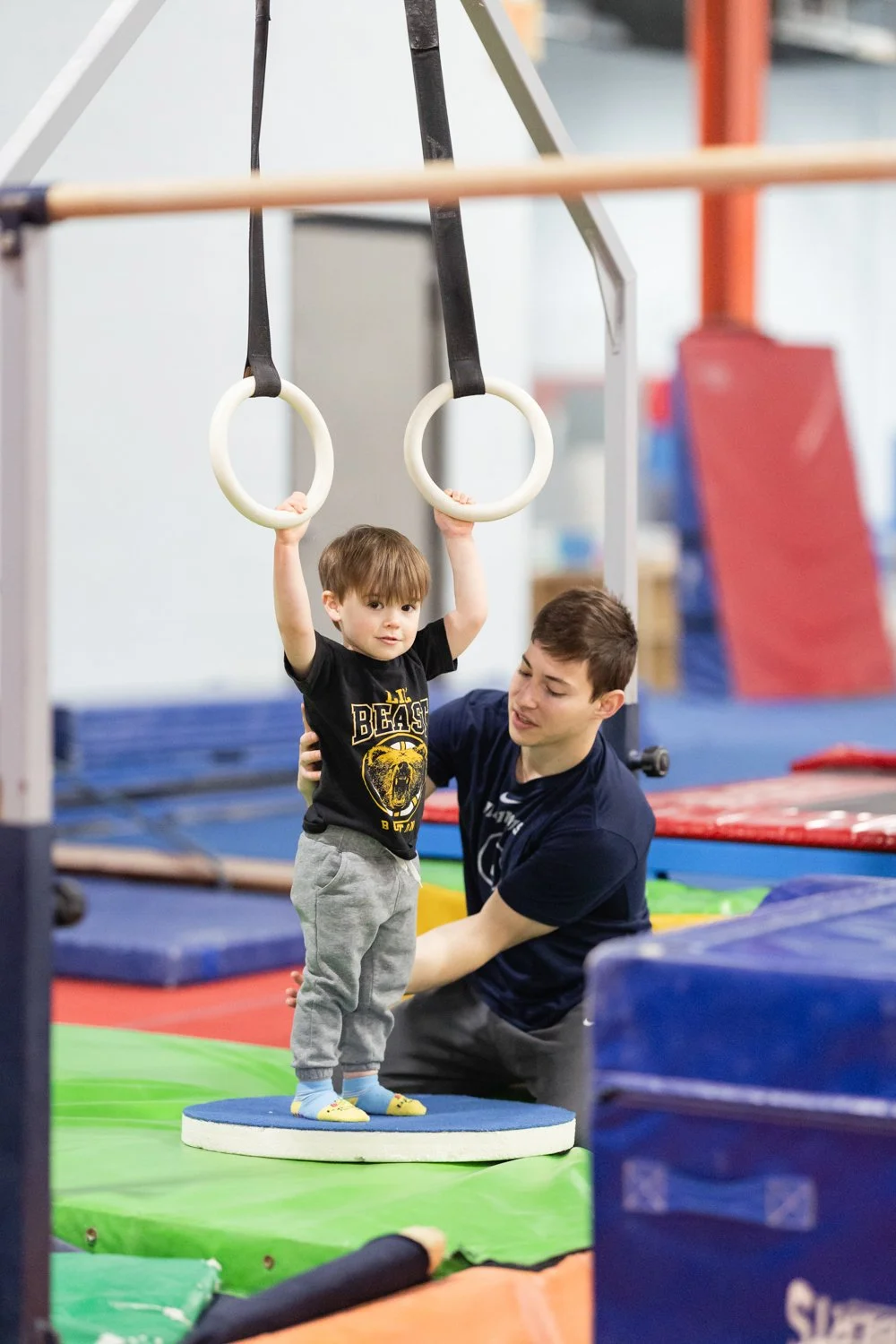 A young boy holding onto gymnastic rings with a trainer supporting him in an indoor gym facility.