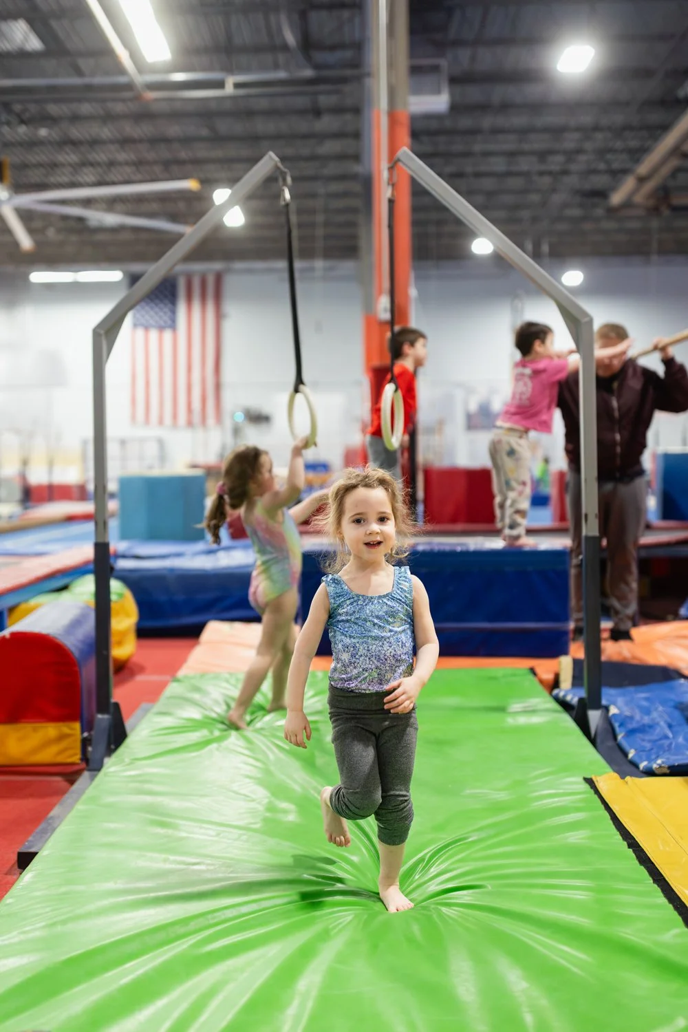 Children playing and running on a padded play area inside a gymnasium with gymnastics equipment and an American flag in the background.