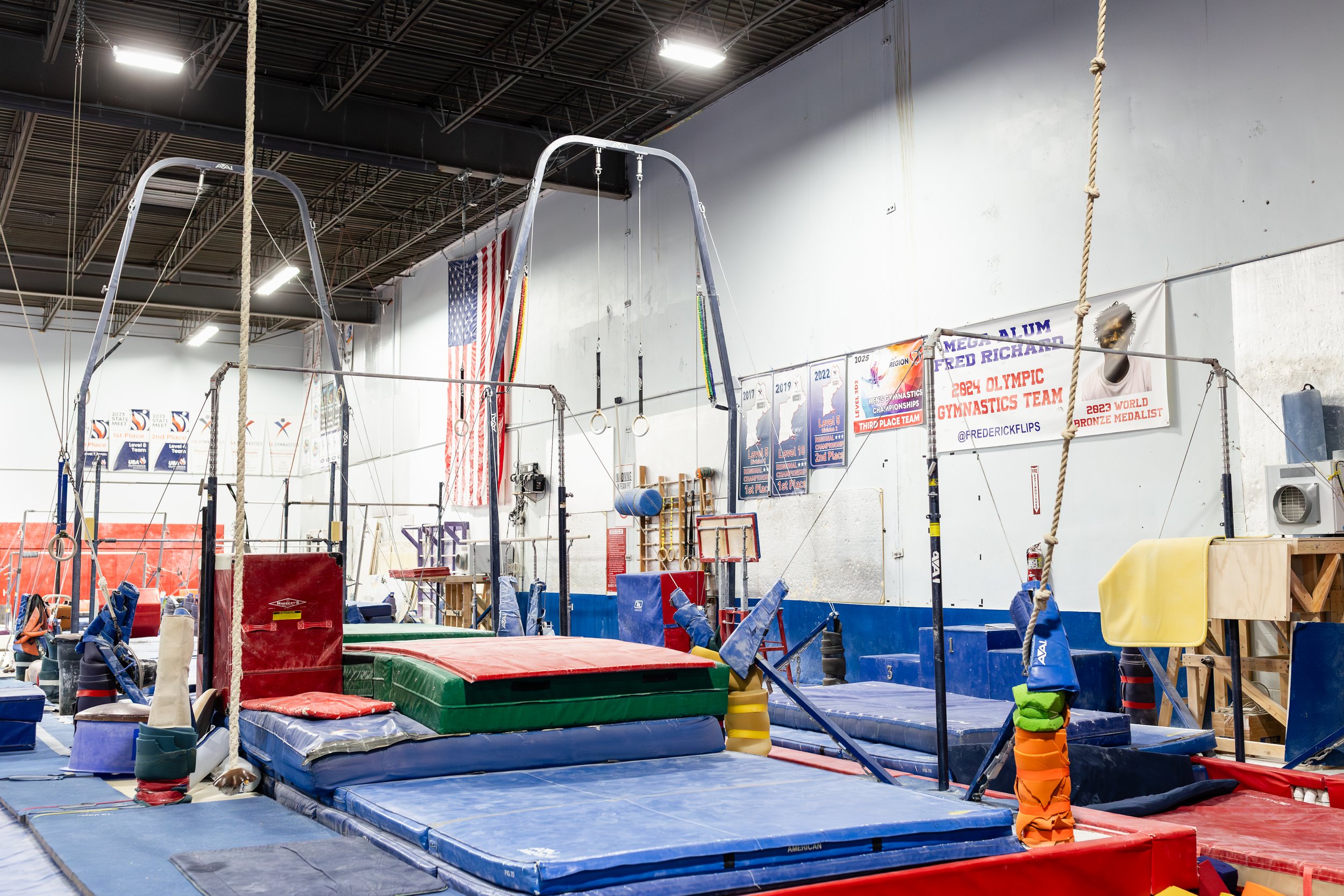 Inside a gymnastics training facility with various mats, gymnastic equipment, and a scoreboard with banners and flags hanging on the wall.