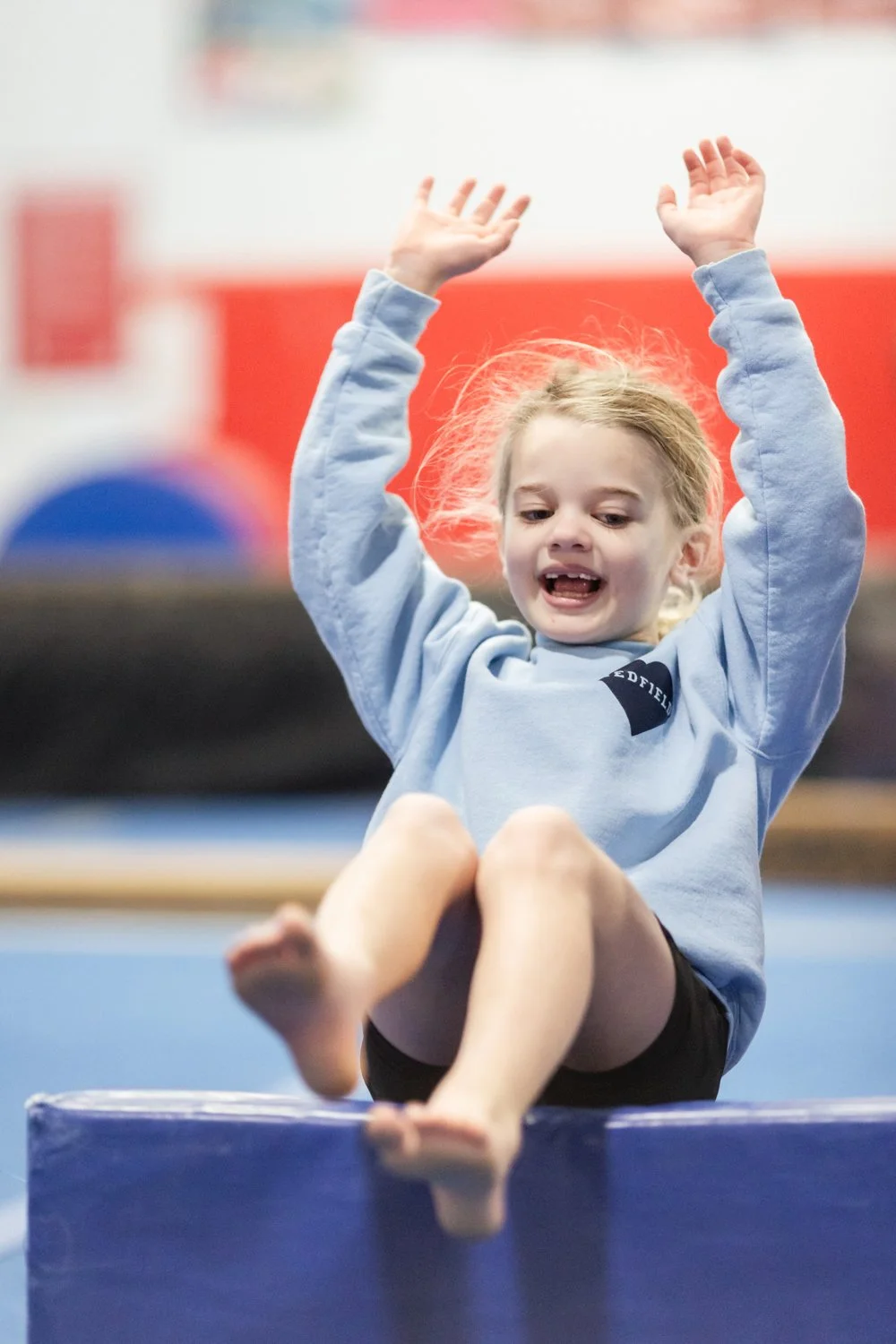 Young girl with blonde hair, wearing a light blue sweatshirt and black shorts, is mid-jump on a gymnastics beam with her arms raised, inside a gymnasium with red and white walls.