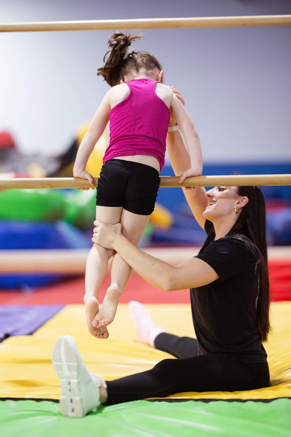 A young girl in a pink tank top and black shorts is holding onto a horizontal bar during a gymnastics session. A woman is sitting on the mat, smiling as she assists the girl with her exercise.