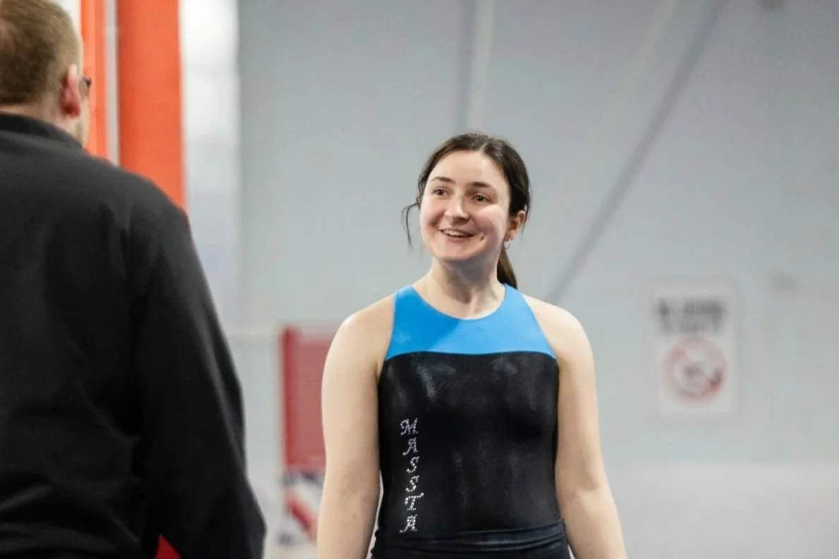 A female gymnast with brown hair wearing a black and blue leotard smiling and talking to a man in a black jacket in an indoor gymnasium.