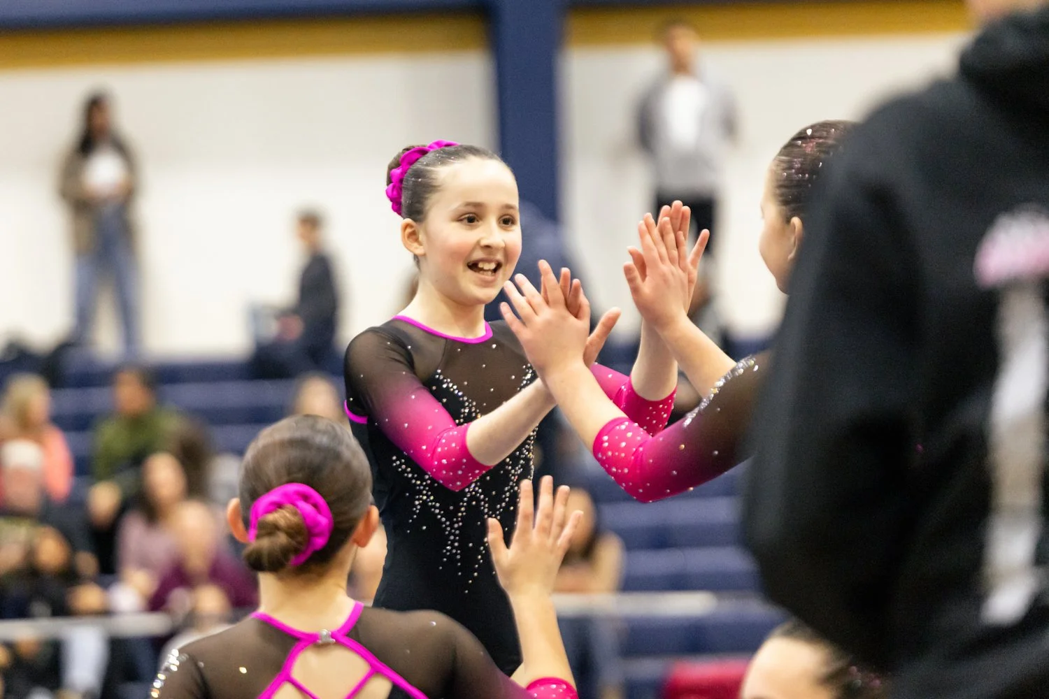 Young girls in gymnastics leotards high-fiving in a gymnasium with spectators in the background.